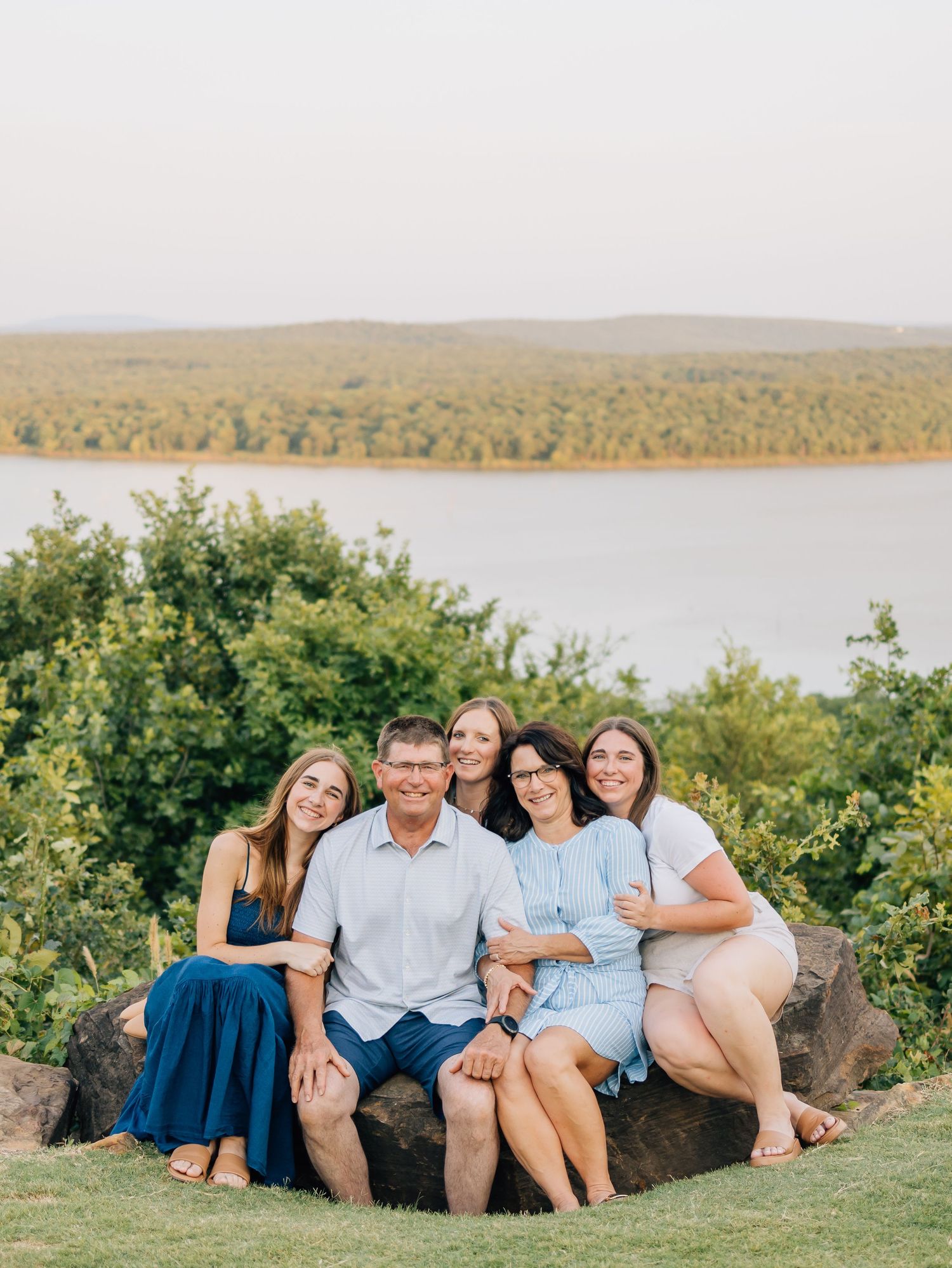 A family group poses together on a rock overlooking a scenic lake and forest landscape.