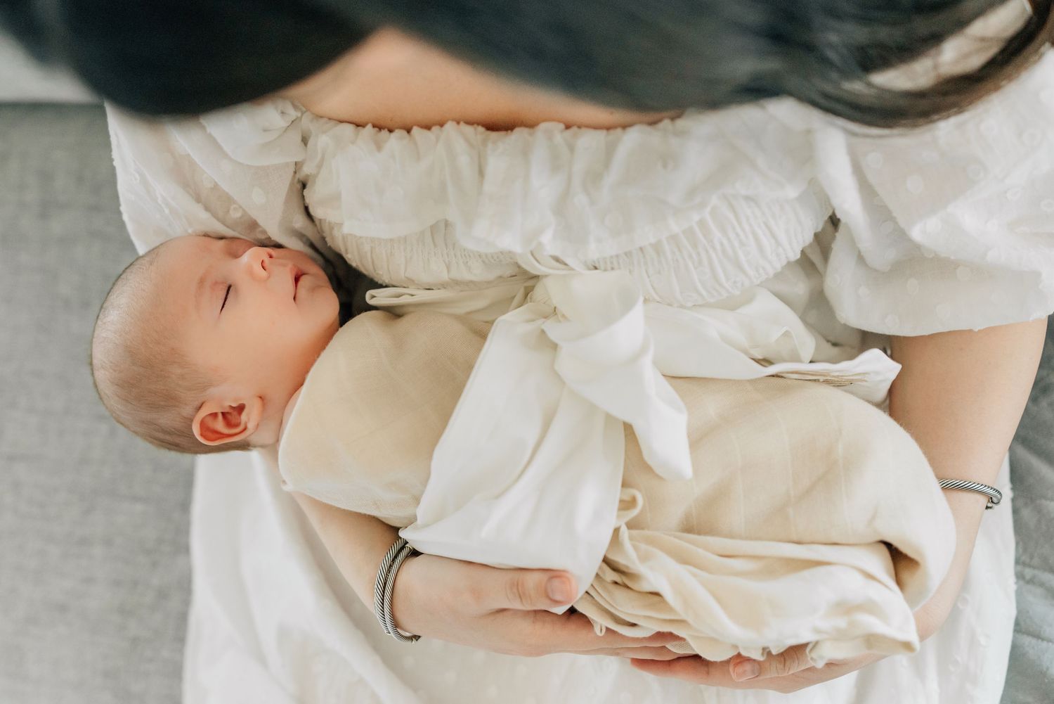 A mother tenderly cradles her sleeping newborn baby wrapped in a soft white swaddle blanket.