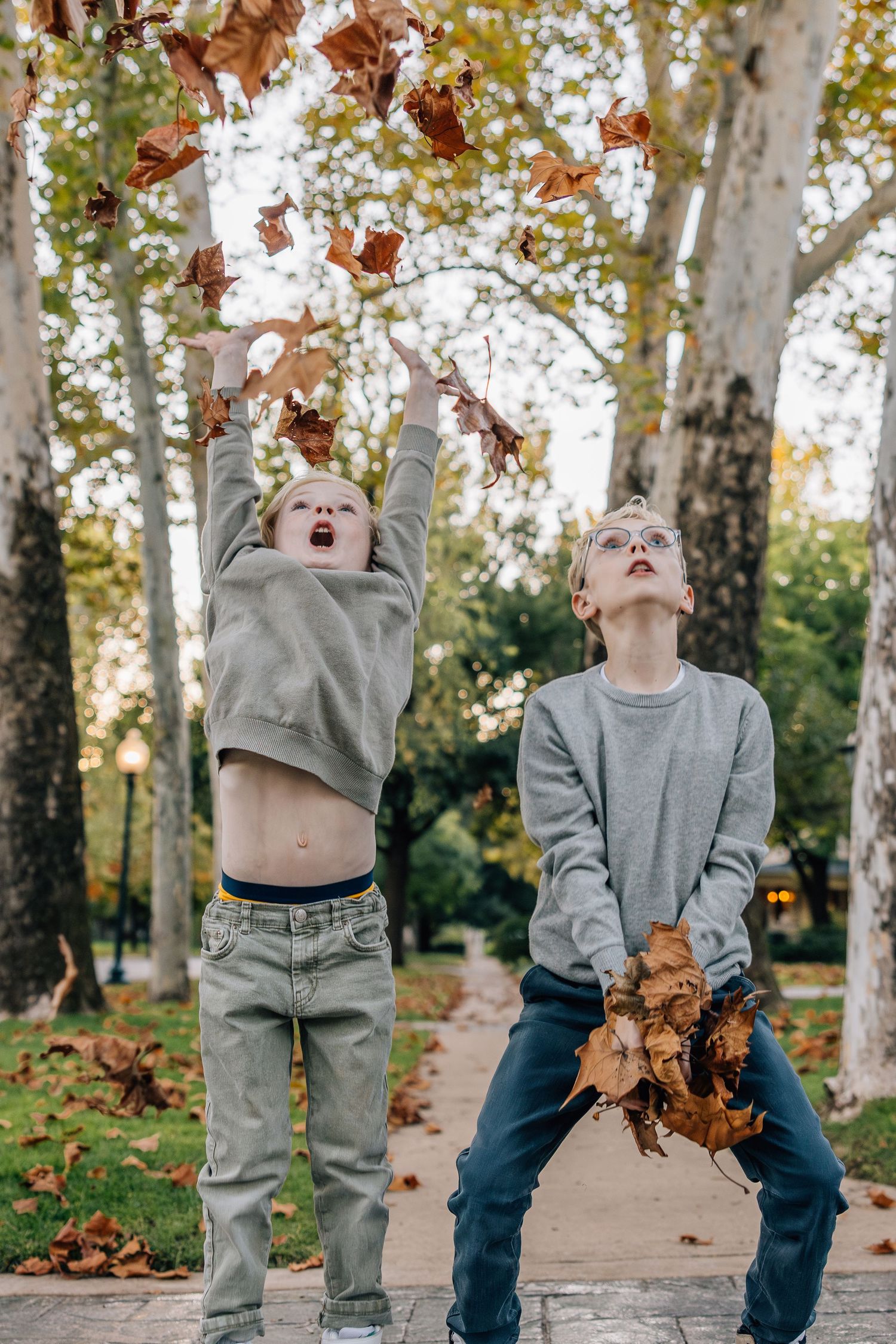 Two kids playing with autumn leaves in a park on a fall day, throwing foliage in the air with joy.