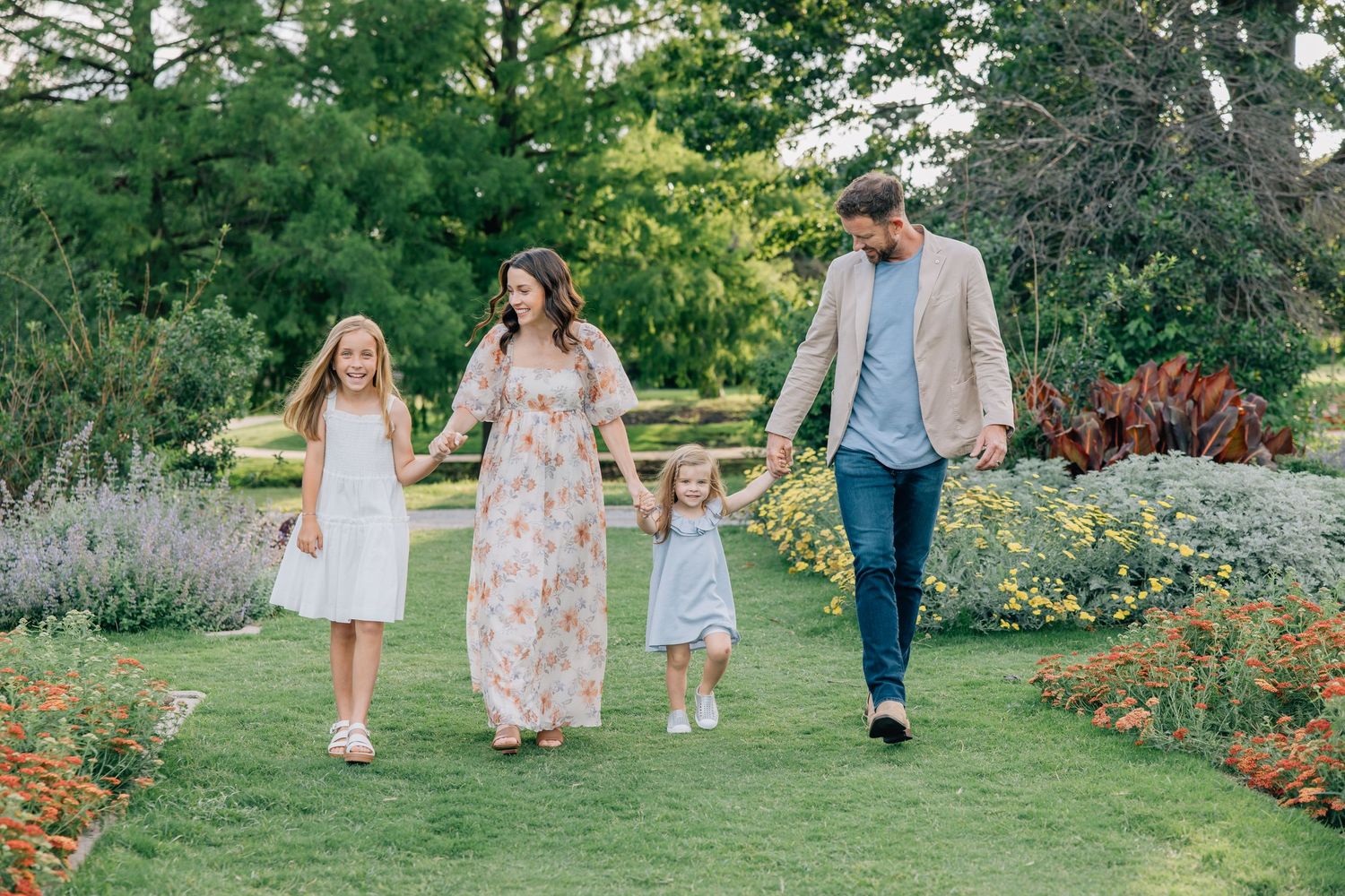 People walking together through lush summer gardens with blooming flowers and green landscaping in a dreamy outdoor setting.
