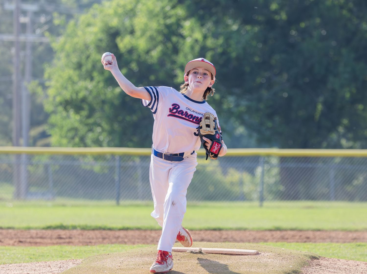 Young baseball player in white uniform follows through on pitching motion during game on sunny day at baseball field.