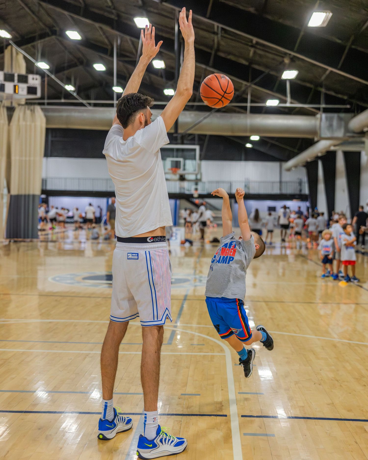 Two basketball players in a gym, one reaching up high while a younger player attempts to shoot the ball.