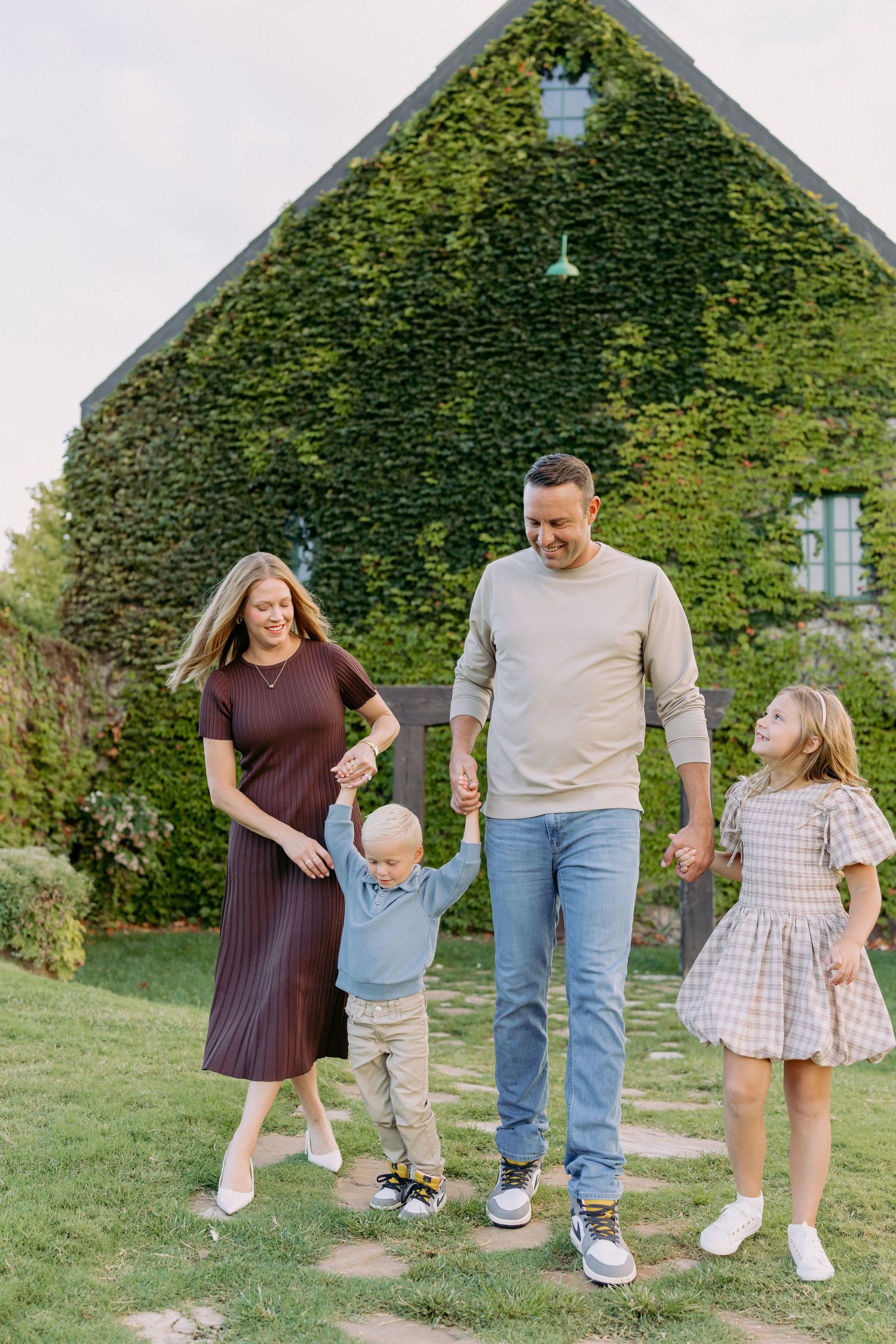 A family walks together on a lawn path in front of an ivy-covered house on a sunny day.