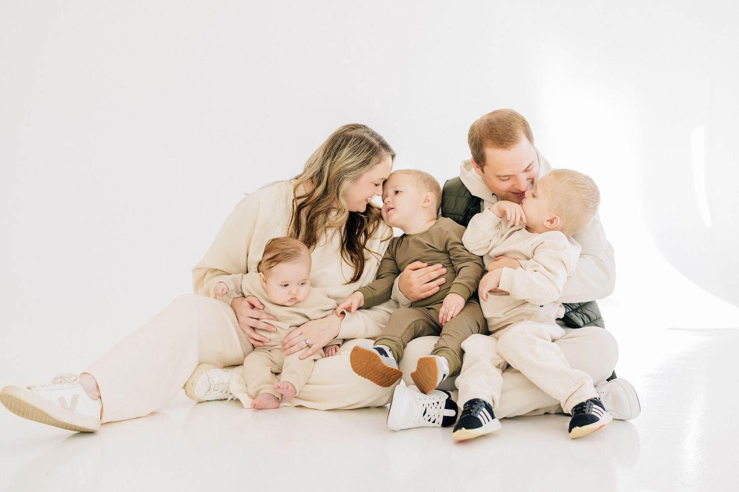 Family in white clothing sits together on floor in bright studio setting sharing affectionate moment.