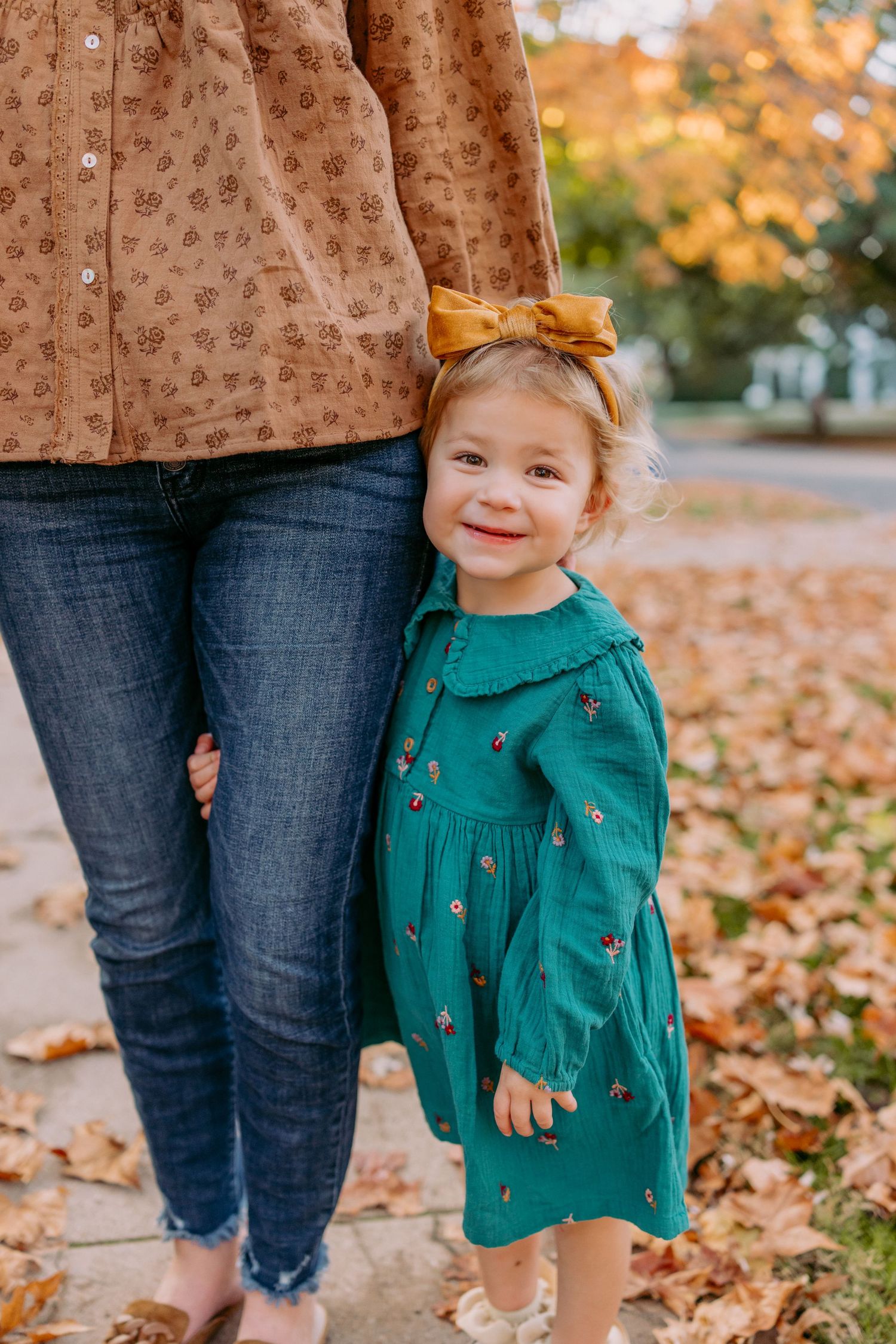 Small child wearing a teal sweater smiles while playing in fallen autumn leaves on a sunny day.