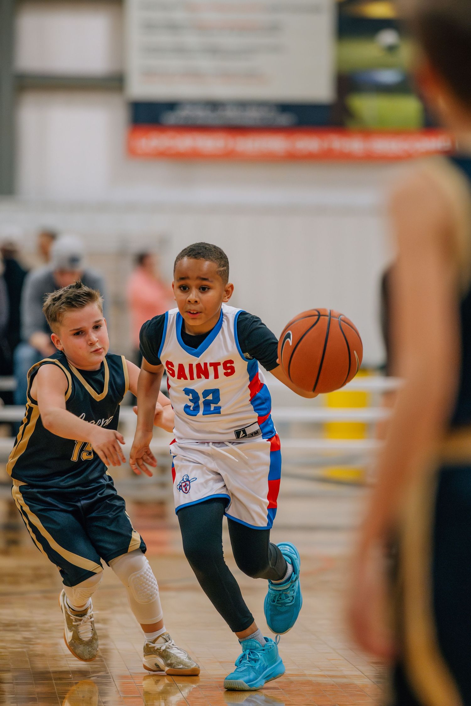 Youth basketball players compete during a game, with one in a blue Saints jersey dribbling past a defender.