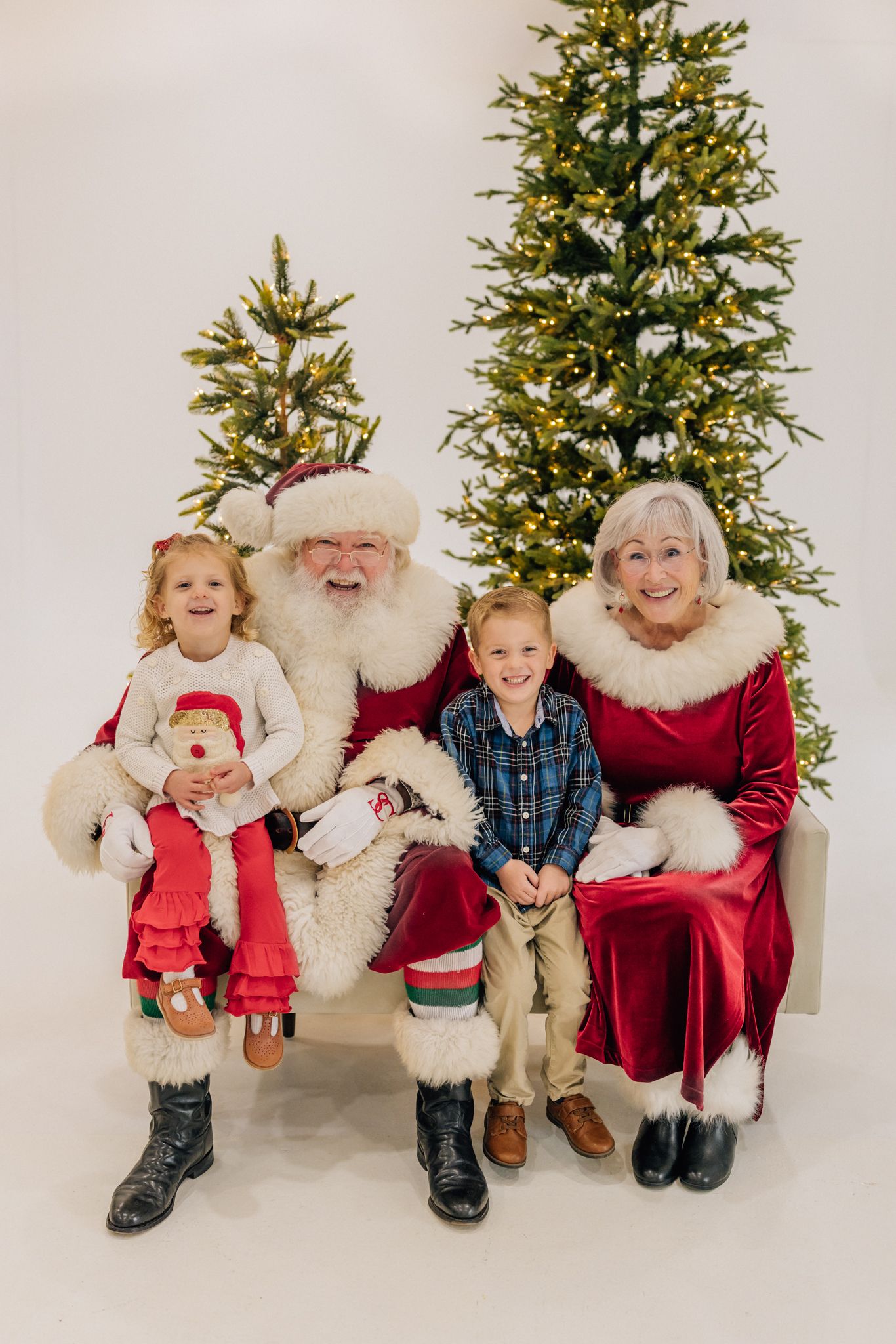 Two children pose with Santa Claus and Mrs. Claus in front of lit Christmas trees in a festive holiday portrait.