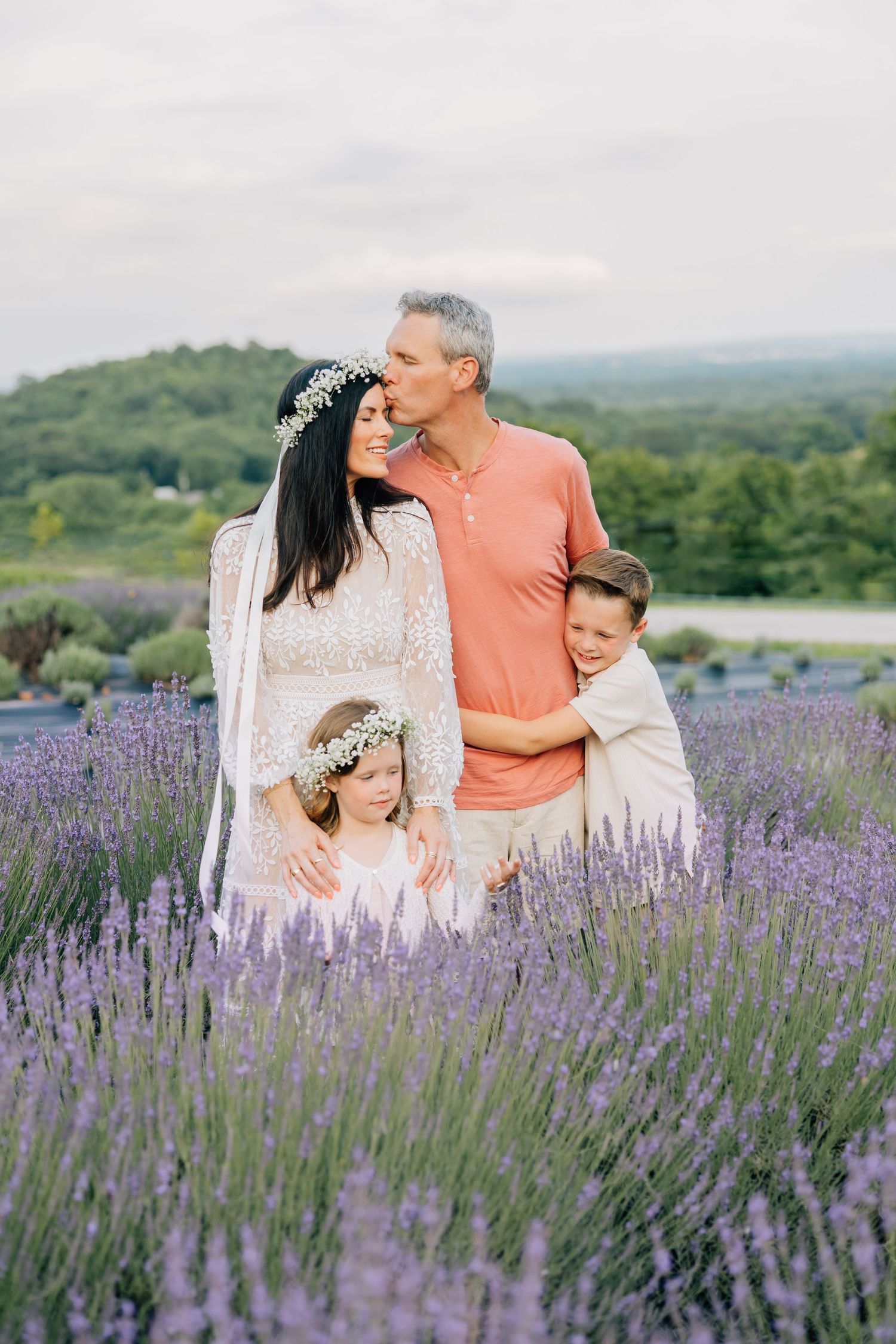 A family wearing white and coral shares a tender moment together in a blooming lavender field on a summer evening.