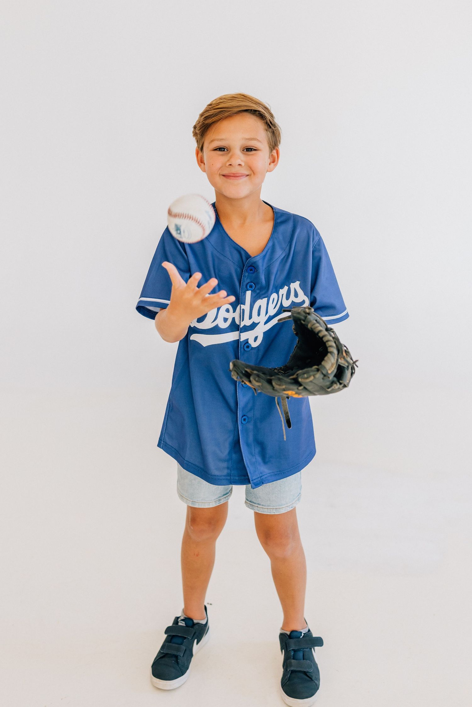 Young baseball player in Dodgers jersey poses with a baseball and glove against white background.