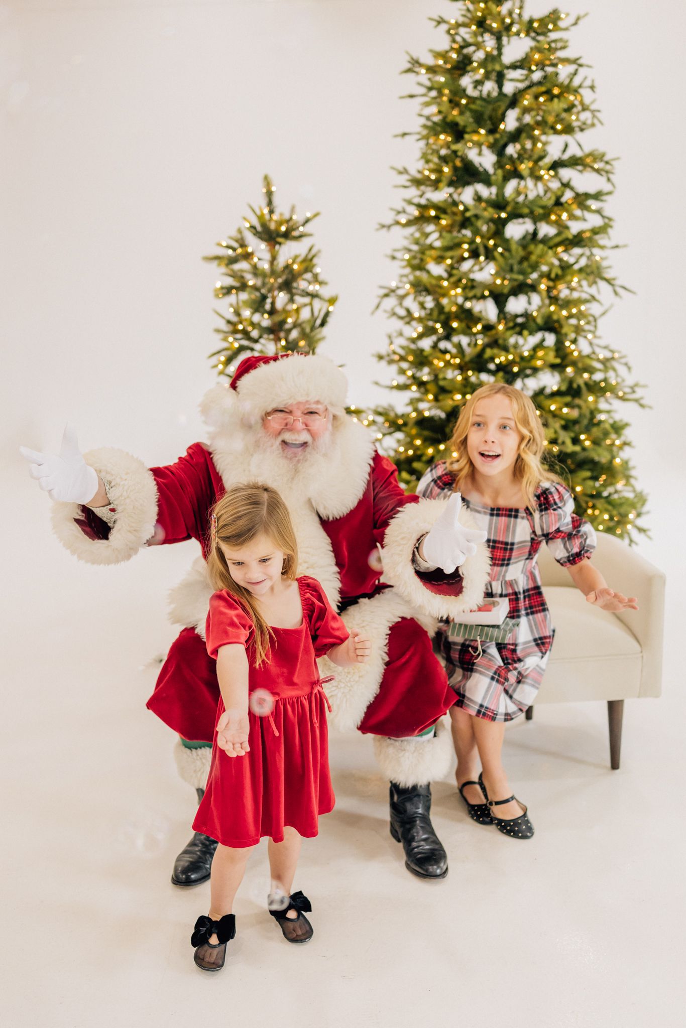 Two girls in holiday dresses pose with Santa Claus in front of a lit Christmas tree in a bright white studio.