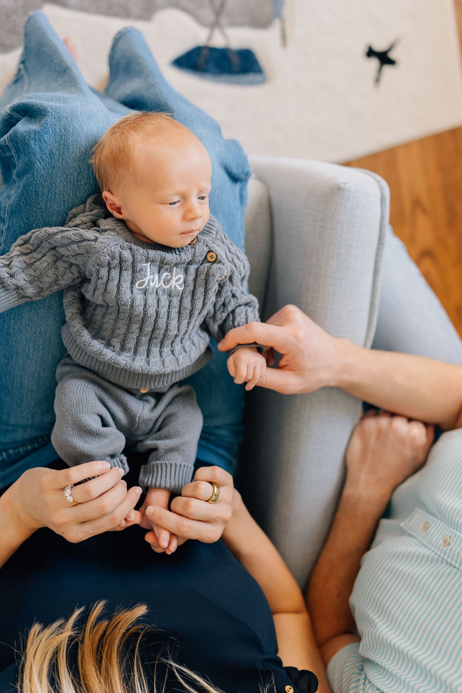 A small child wearing a gray knit sweater sits on a couch while being supported by adult hands.