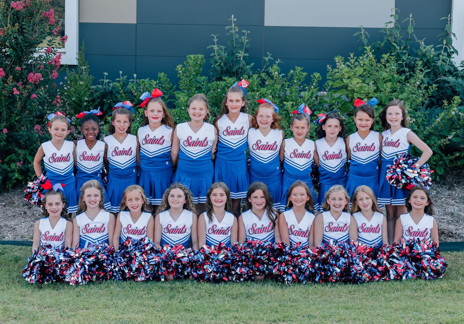 A cheerleading squad wearing white and blue uniforms with pom poms poses together for a group photo outdoors.