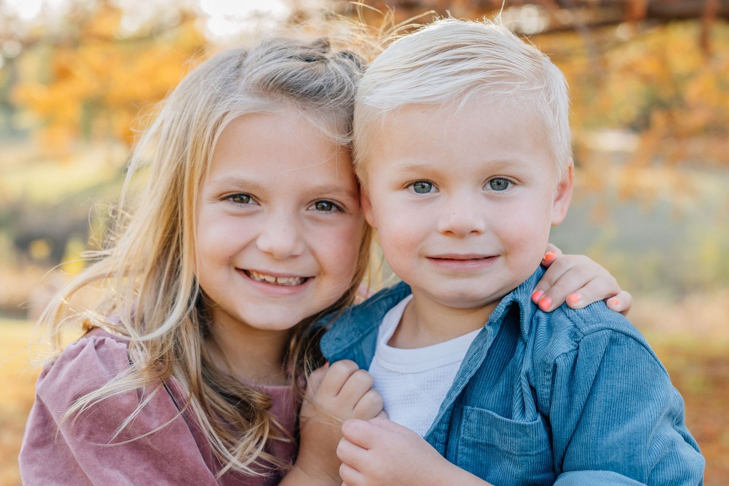 Two young children smile warmly at the camera while embracing outdoors during autumn.
