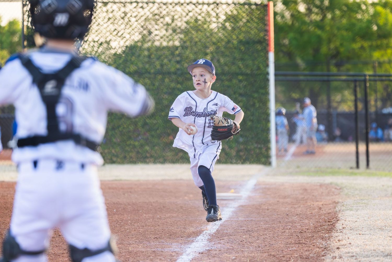 Young baseball pitcher in grey uniform stands ready to throw the ball to catcher on dirt field during youth game.