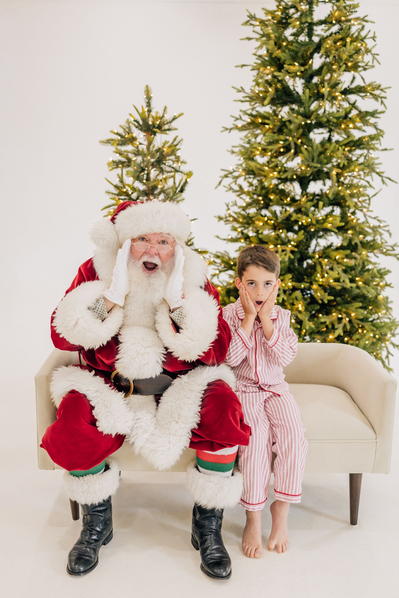 Santa Claus and a child in striped pajamas sit on a couch, both looking surprised, with Christmas trees behind them.