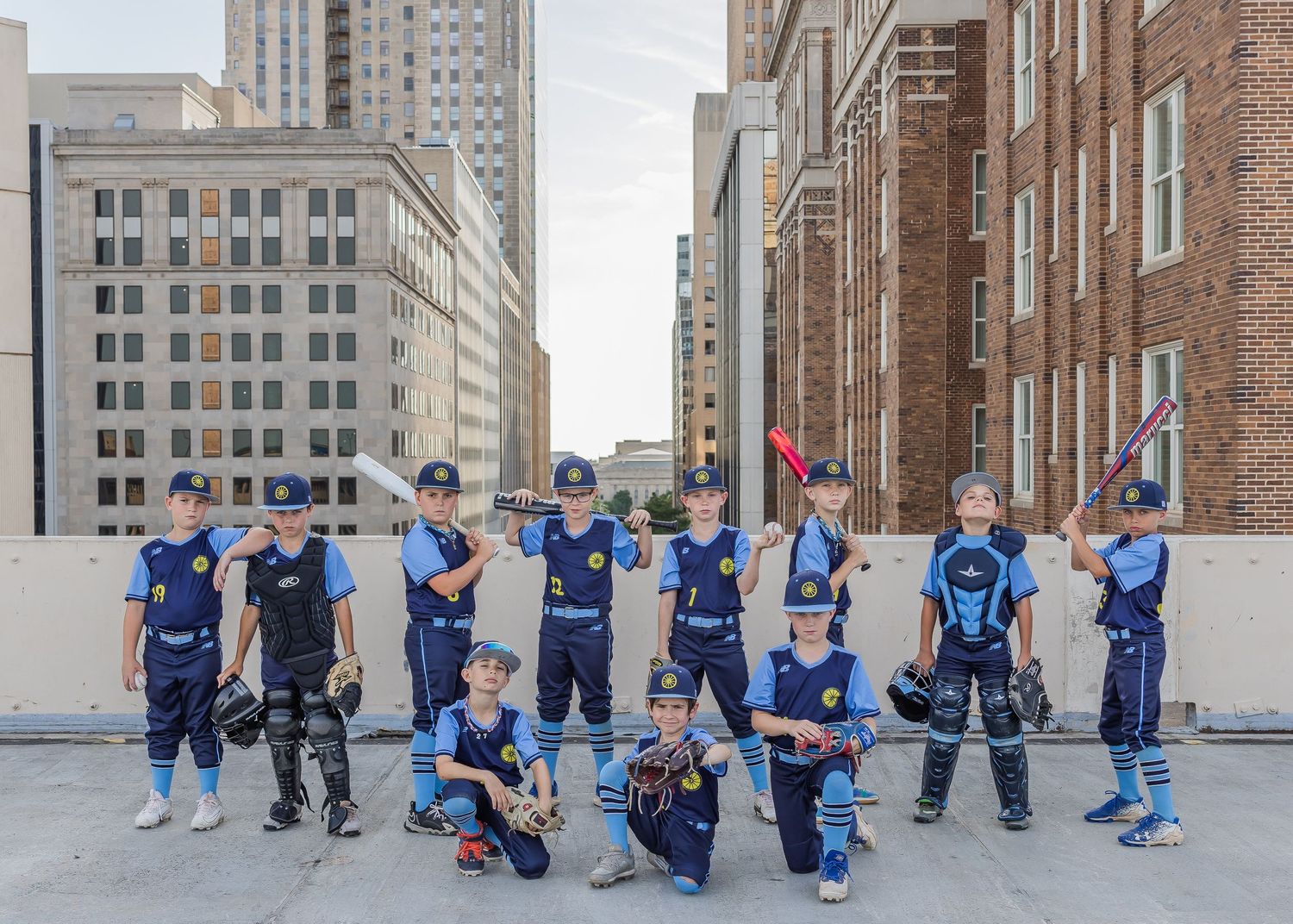 Youth baseball team in matching blue uniforms poses together on an urban rooftop with city buildings in background.