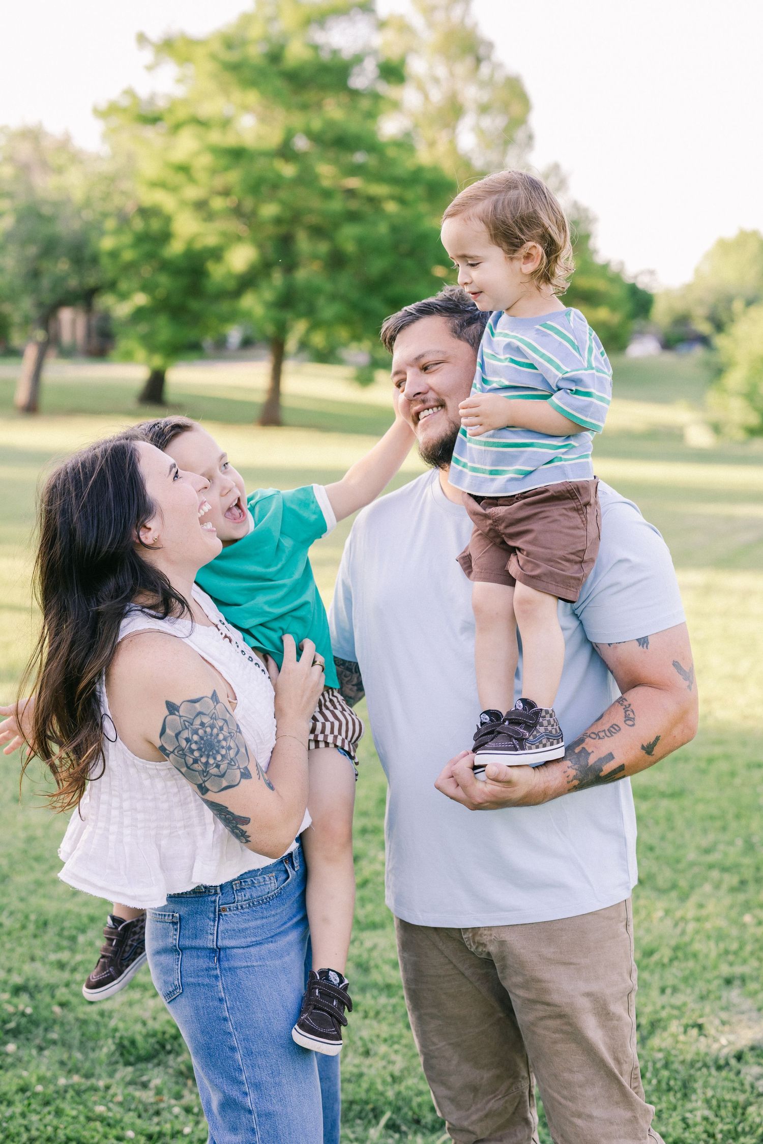 A tattooed family laughing together outdoors, with dad holding a toddler on his shoulders and mom carrying another child.