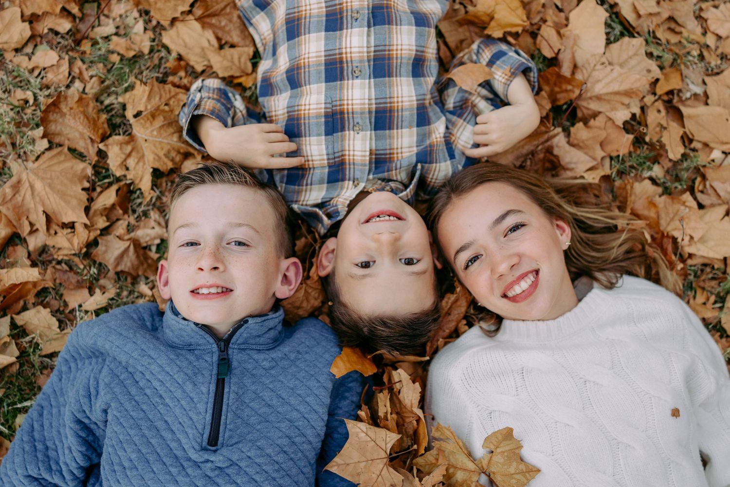 Children laying in autumn leaves wearing blue and plaid clothing while making silly expressions.