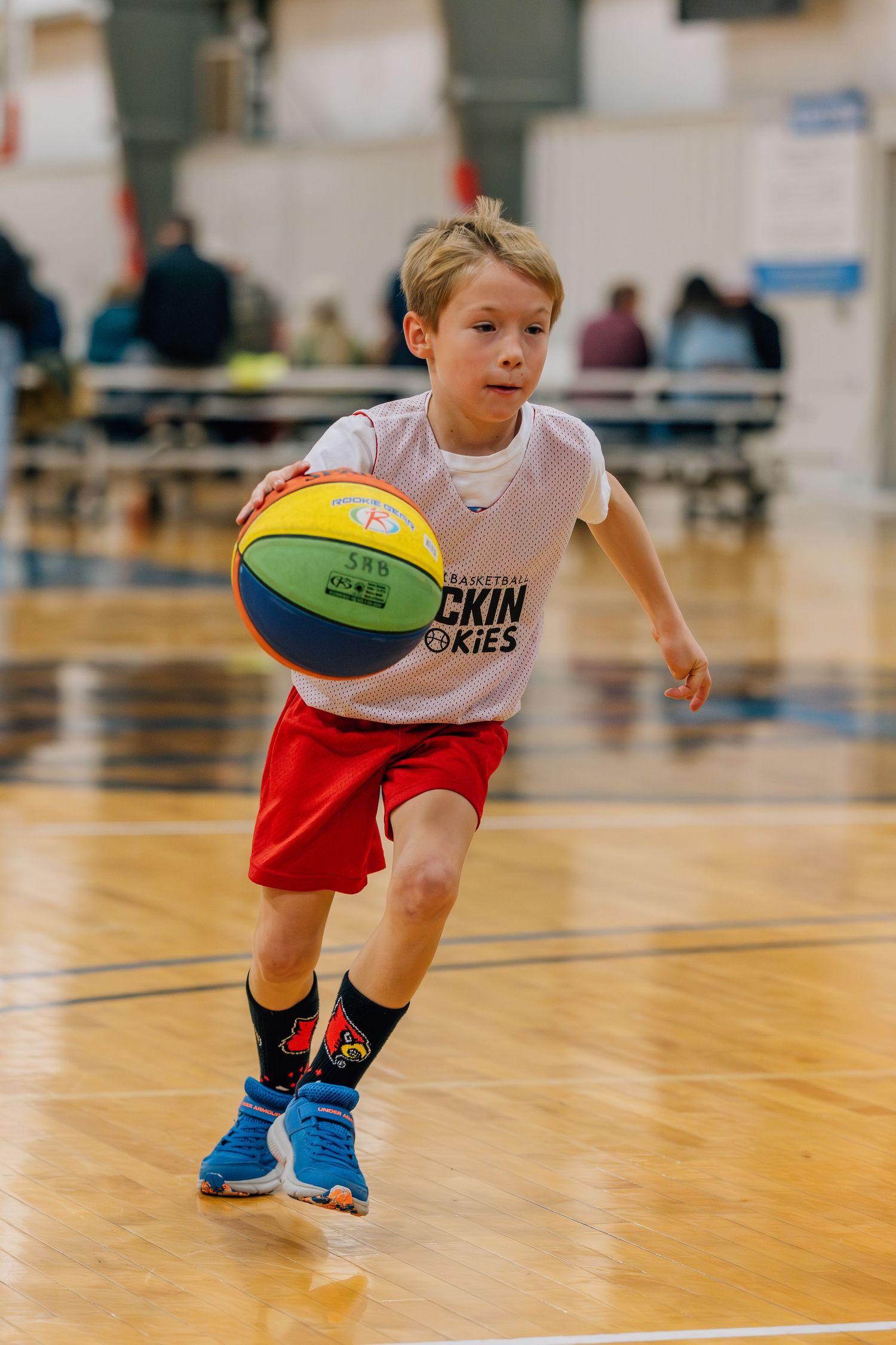 Young athlete dribbles colorful basketball on indoor court while wearing white shirt and red shorts during practice session.