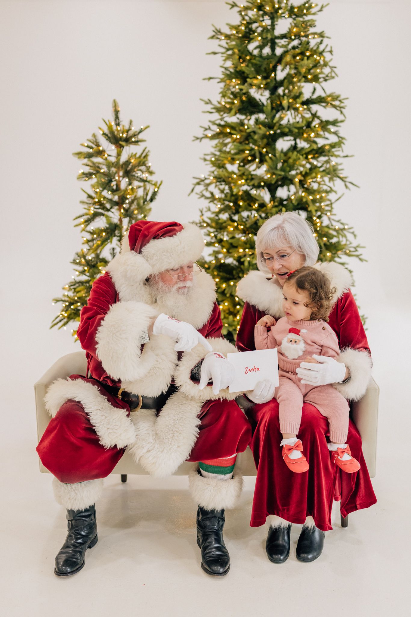 Santa and Mrs. Claus sit with a baby girl on a bench, reading a letter near lit Christmas trees.