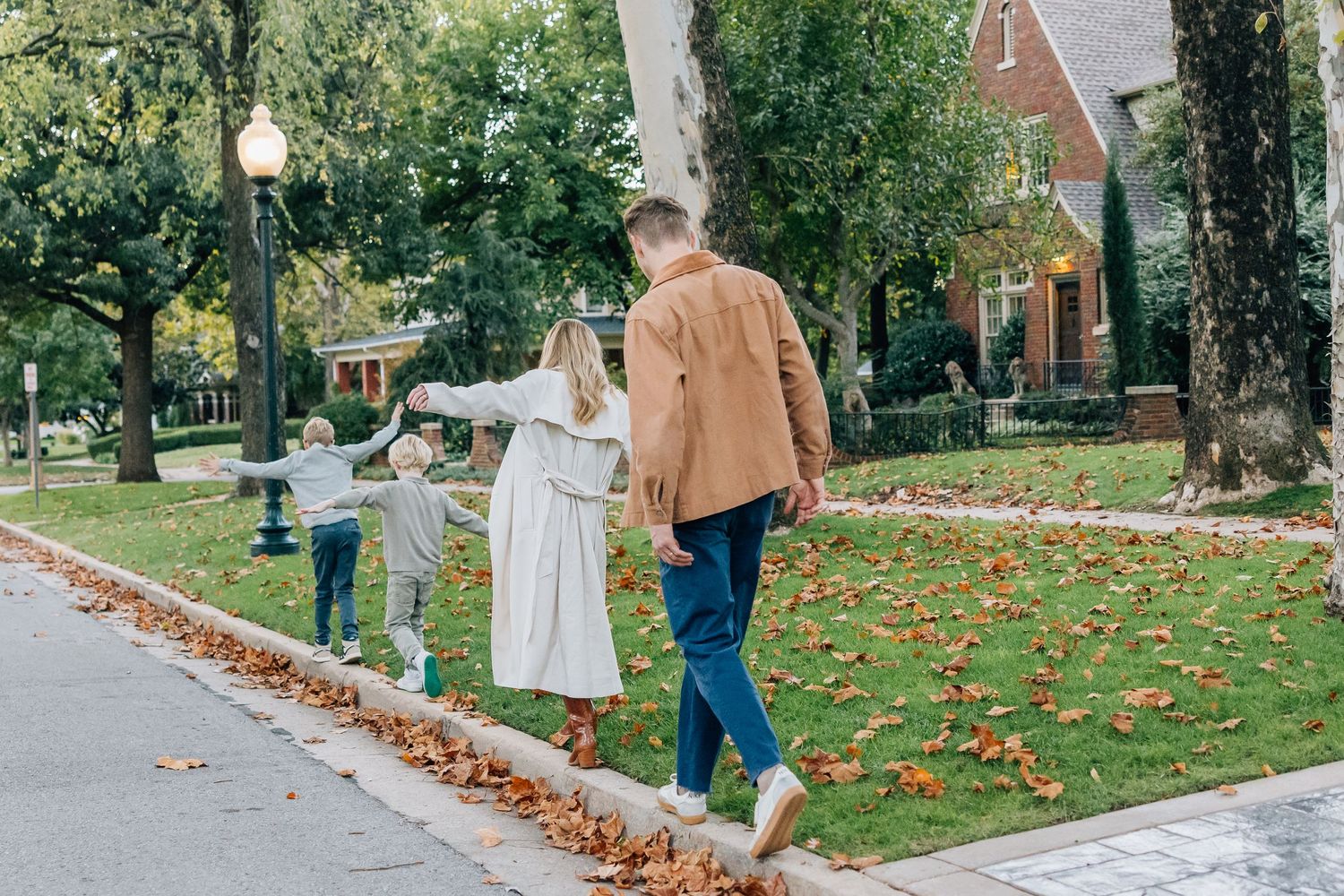 A couple walks hand in hand on an autumn path with fallen leaves and vintage-style buildings in the background.