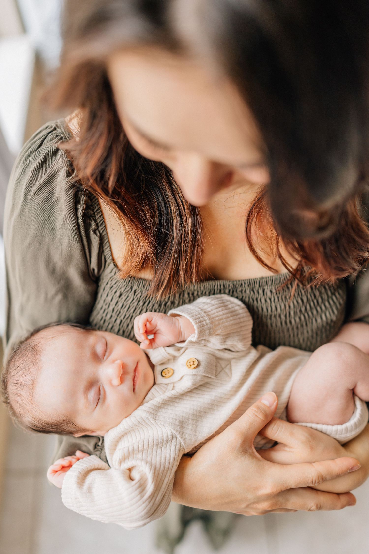 A peacefully sleeping newborn baby in a cream sweater is cradled in loving arms against a soft gray dress.