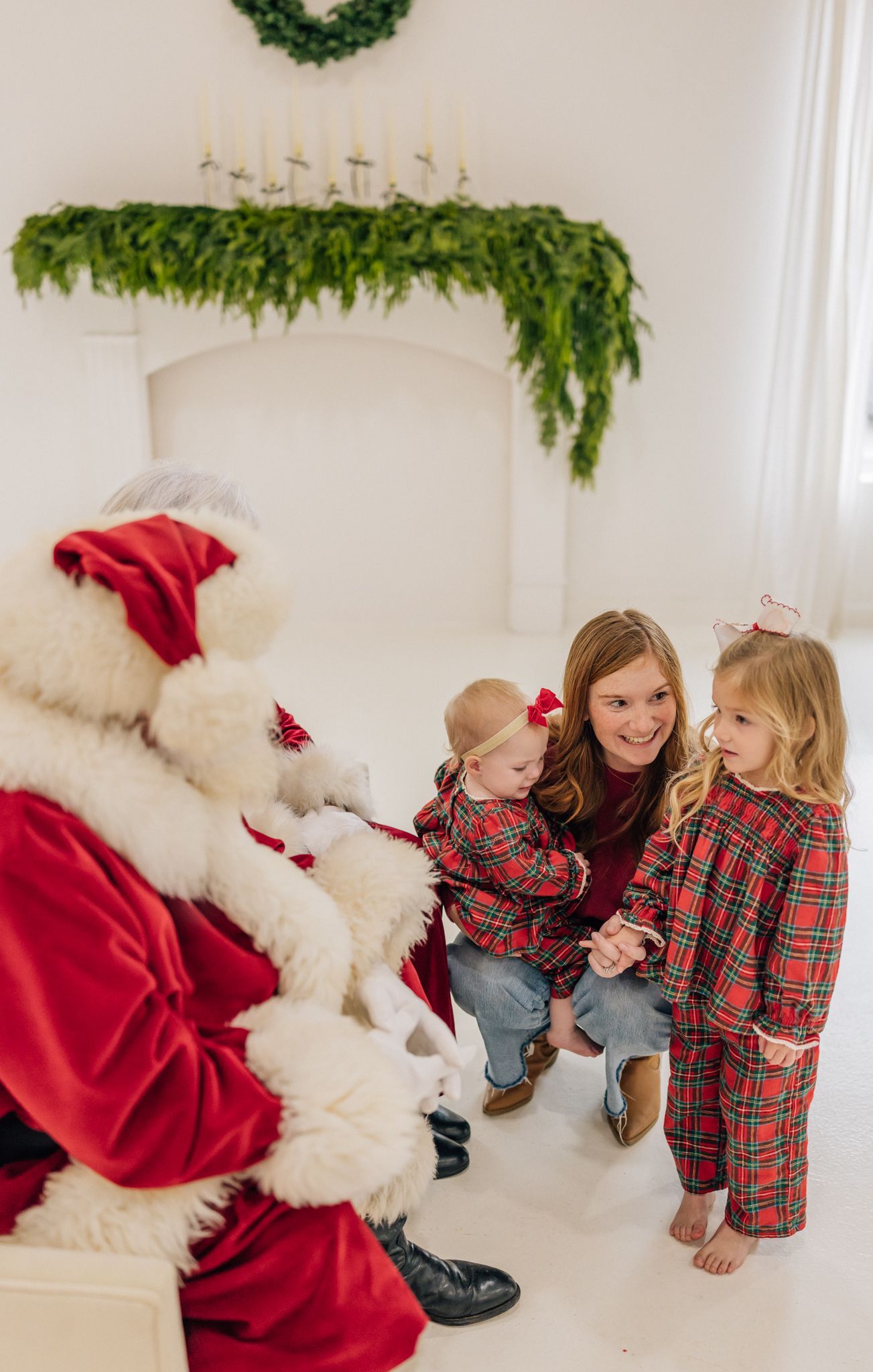 Children in plaid pajamas meet Santa Claus in a white room decorated with festive greenery on a mantel.