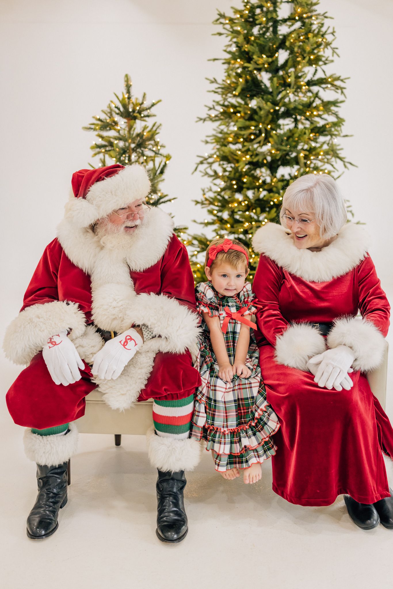 A toddler girl in plaid dress sits between Santa Claus and Mrs. Claus in front of a decorated Christmas tree.