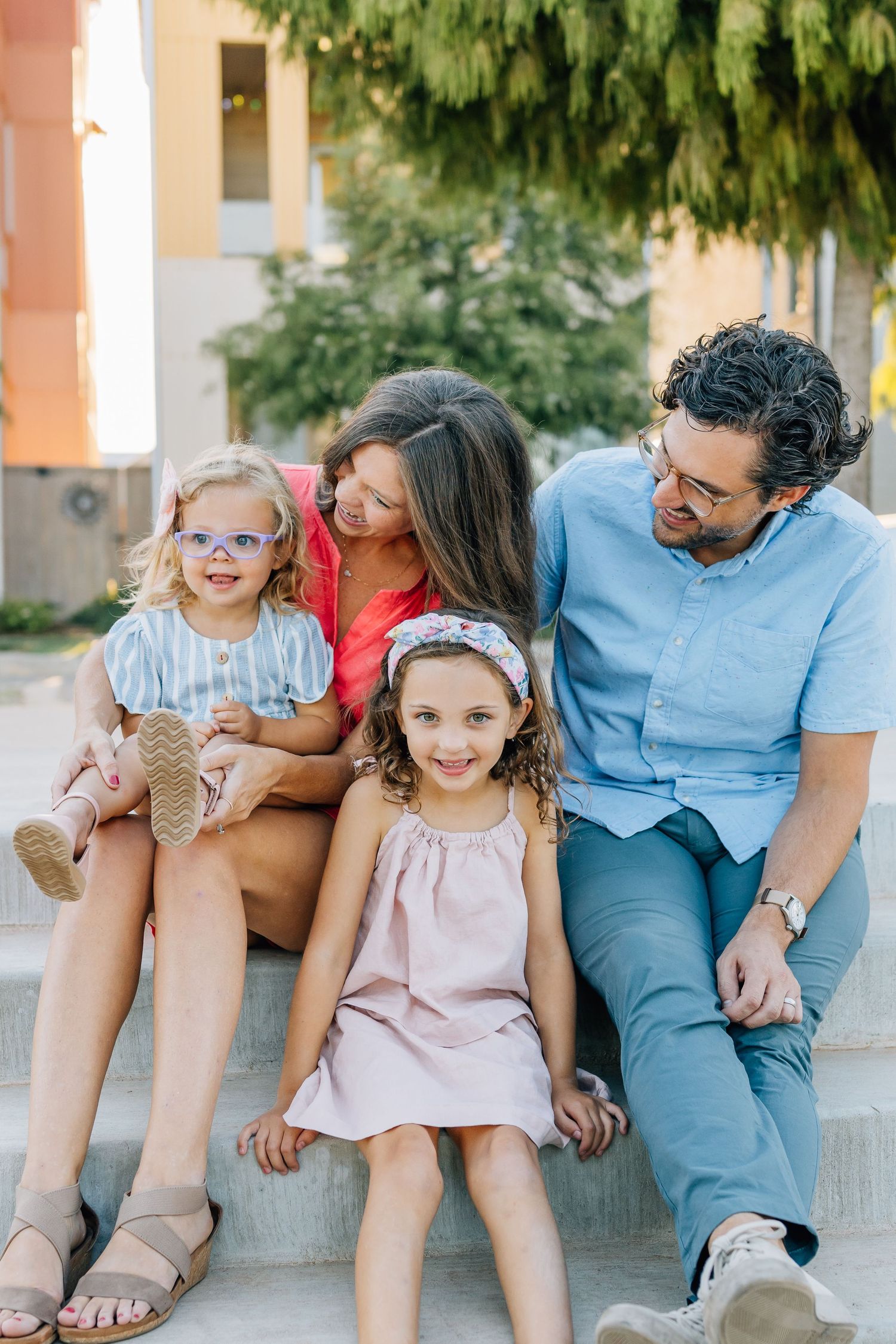 A family of four sits together outdoors on concrete steps, dressed in casual clothing on a sunny day.