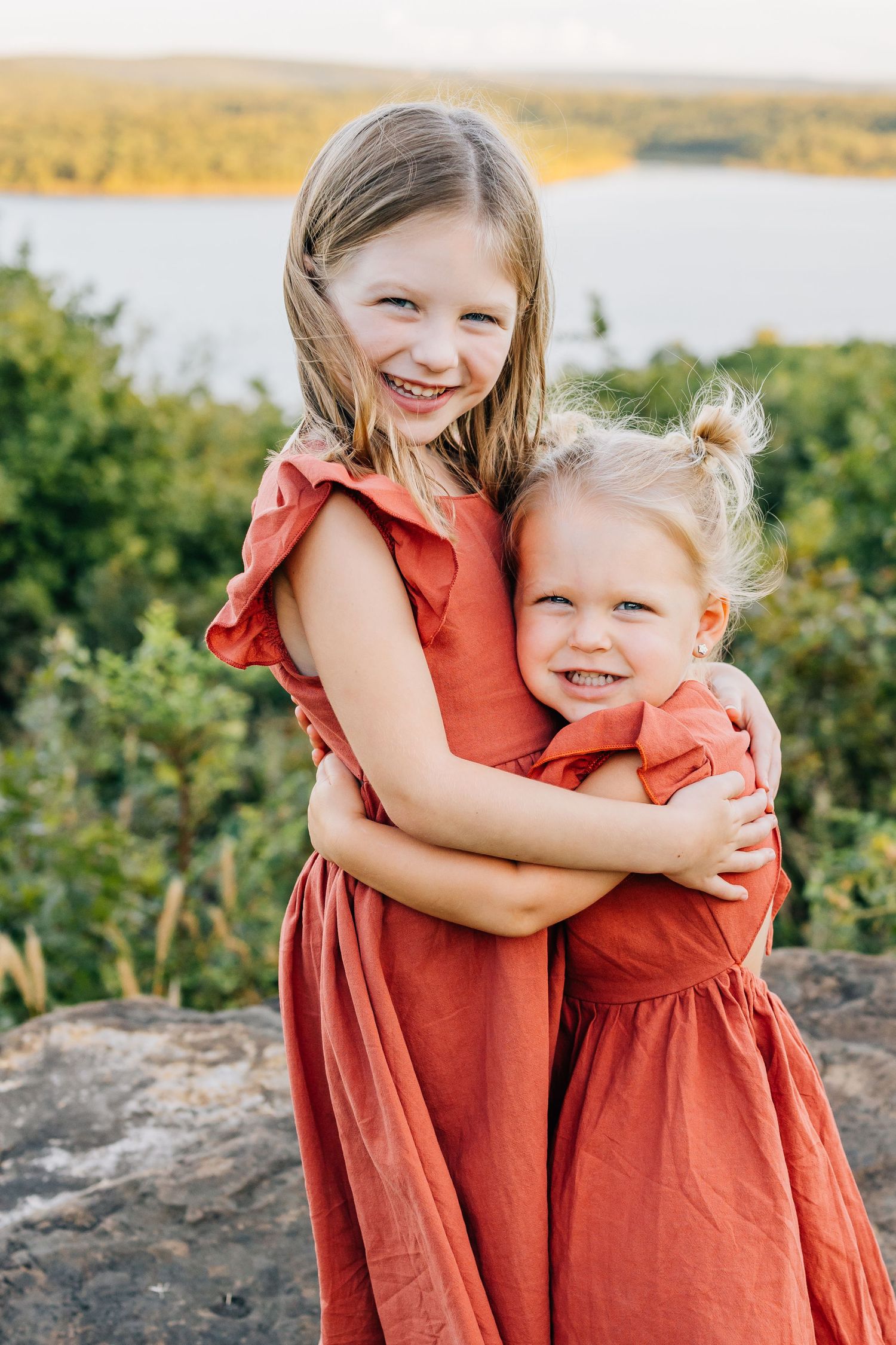 Two children in matching coral dresses hugging and smiling outdoors by a lake.
