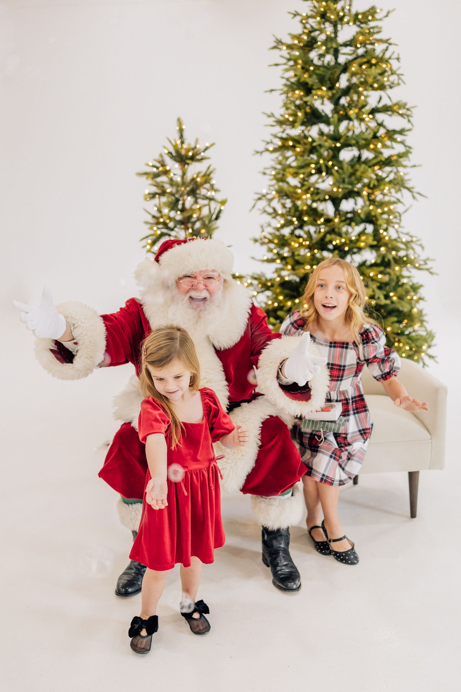 Two girls in festive dresses pose with Santa Claus beside a lit Christmas tree in a bright white studio setting.