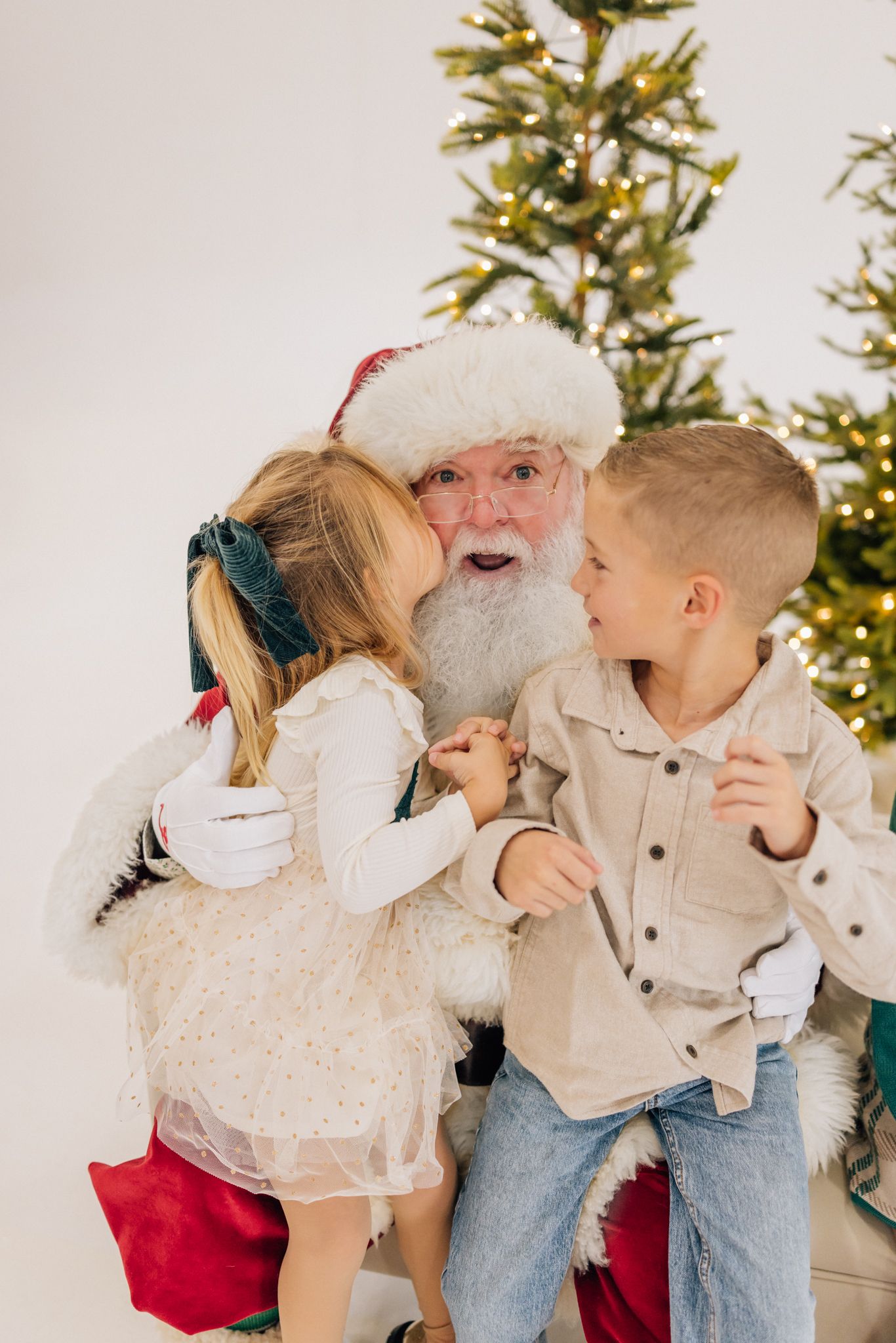 Two children whisper and talk to Santa Claus beside a glowing Christmas tree in a festive holiday portrait.