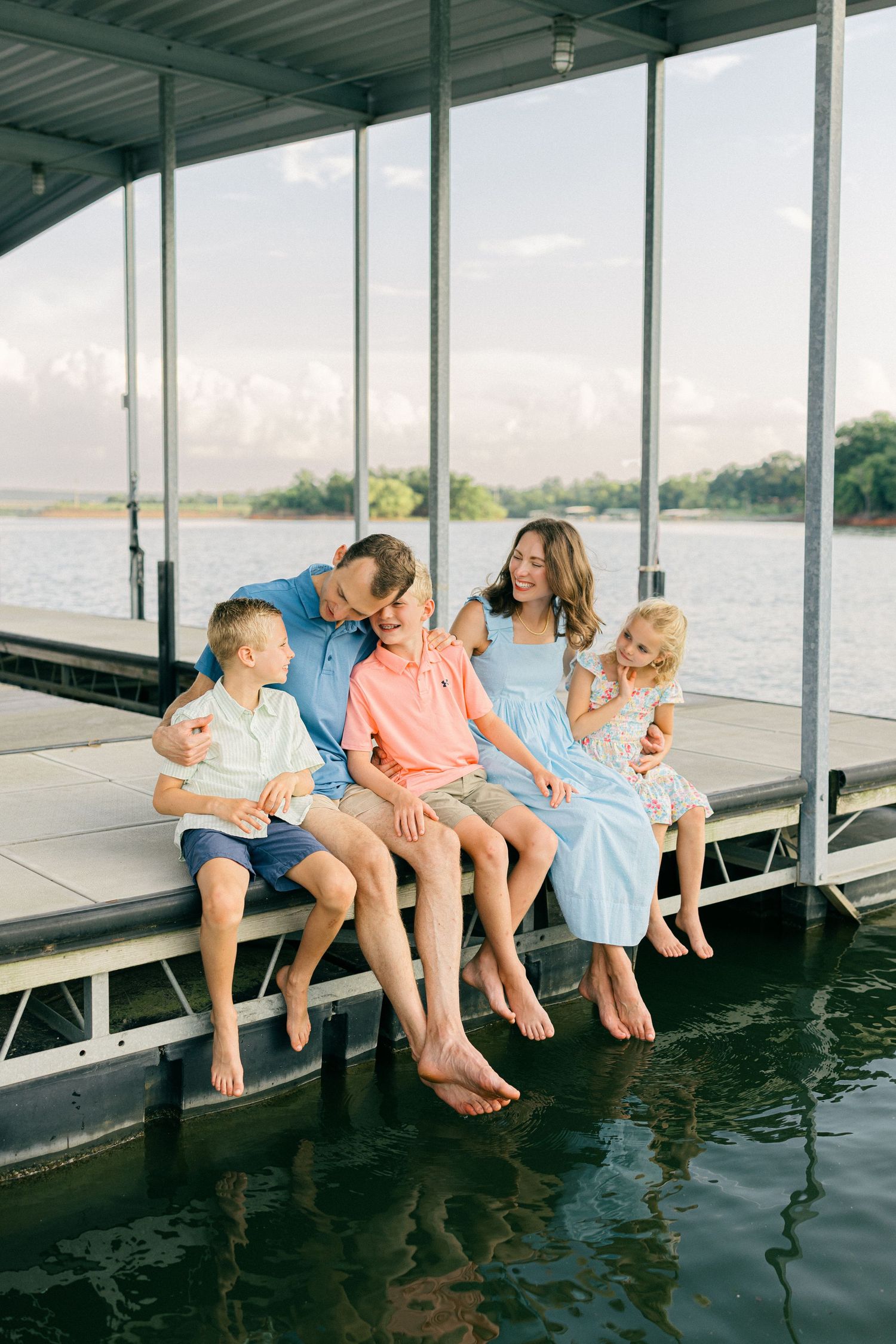 Family of five sitting barefoot on a dock edge over water, smiling and laughing together on a sunny day.