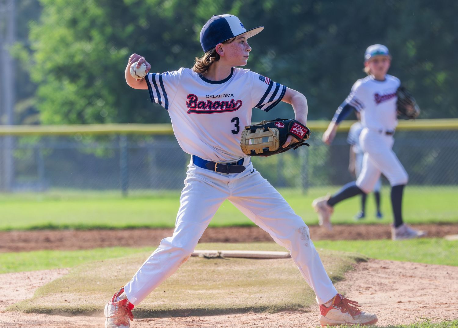 Baseball pitcher in white uniform winds up to throw from the mound during a youth baseball game.