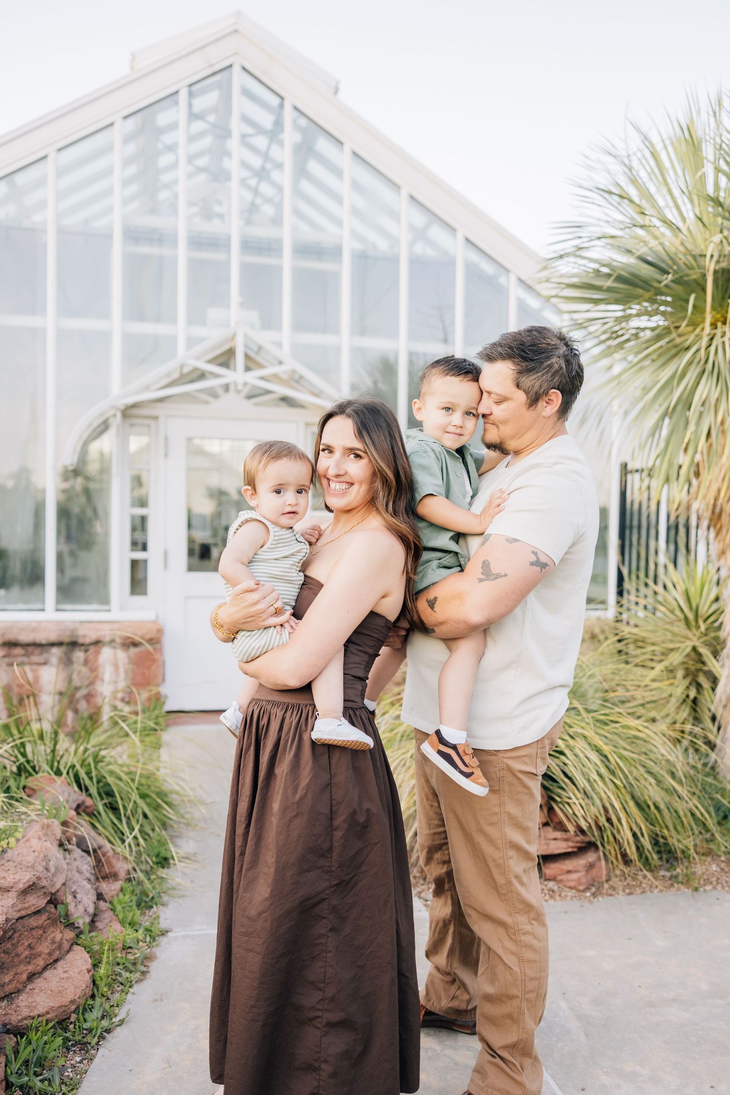 A family poses together outdoors in front of a large glass greenhouse with palm trees in the background.
