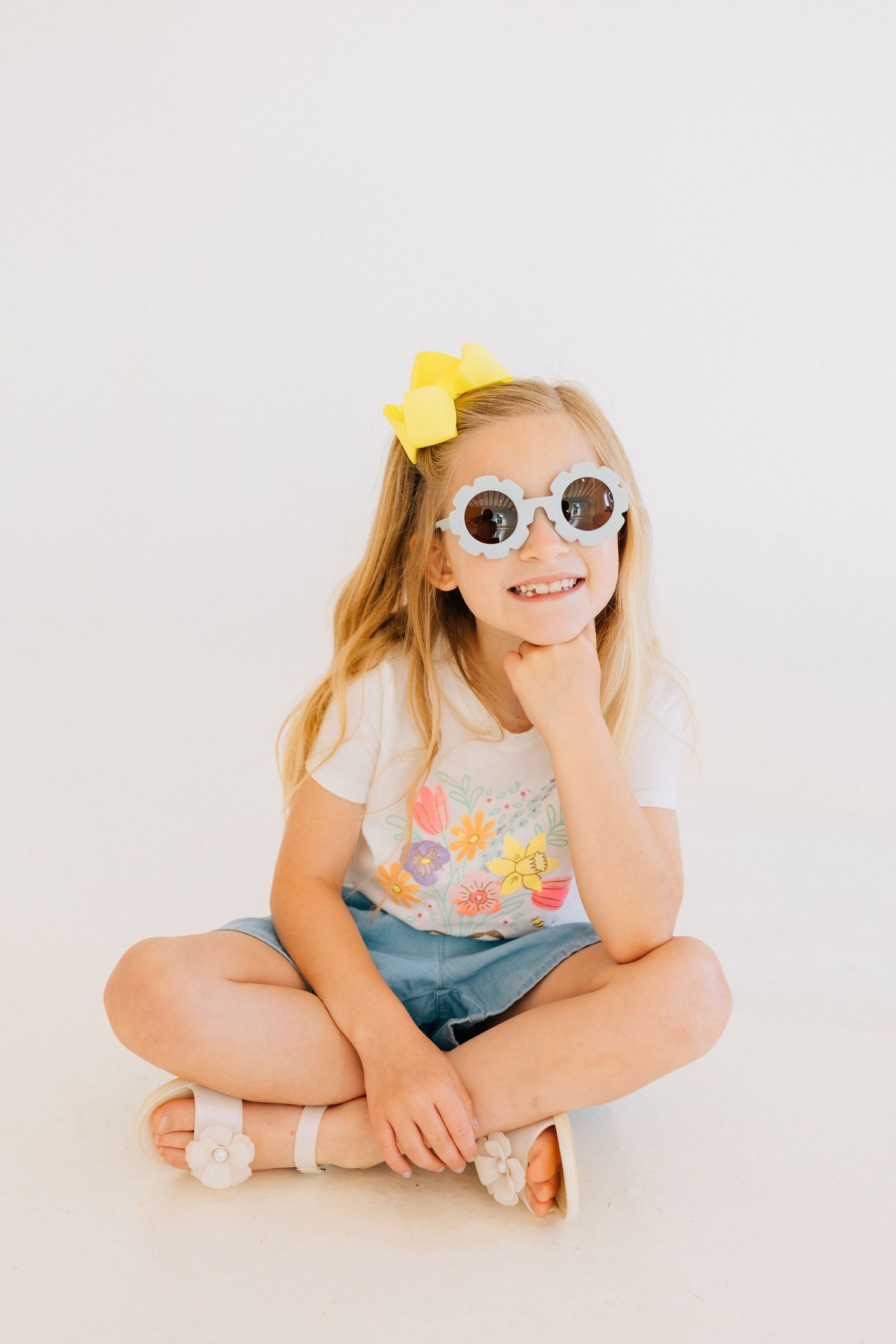 Child in white sunglasses and yellow bow laying on floor in playful poses against light background.