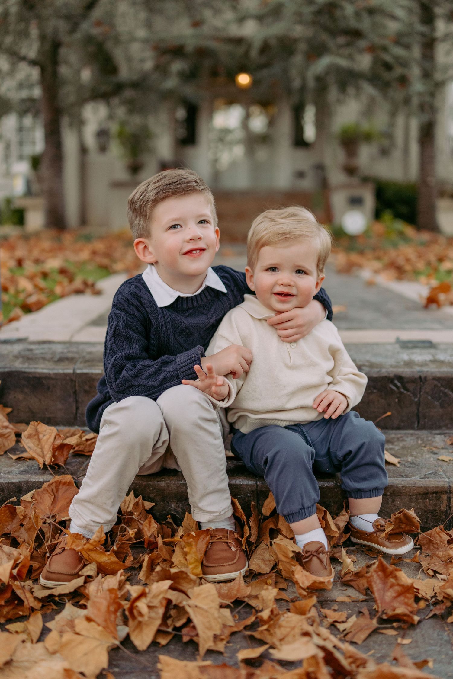 Two young children sit together on stone steps surrounded by fallen autumn leaves in matching brown shoes.