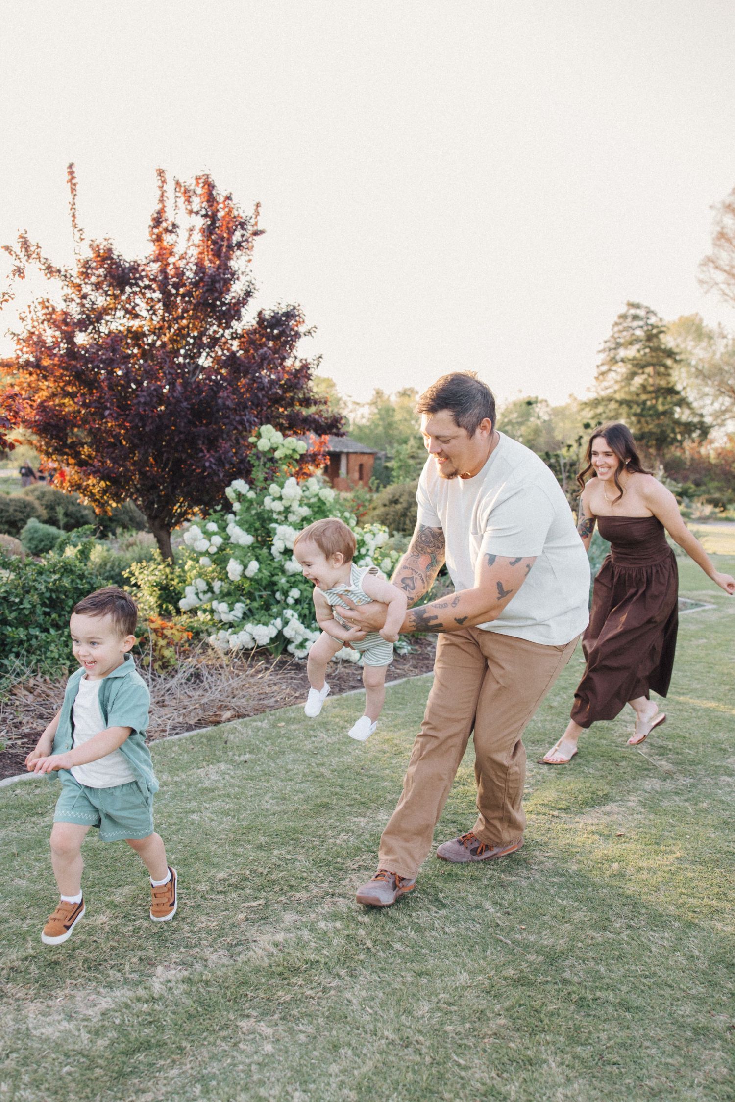 A happy family plays and runs together on green grass during a warm sunset with fall foliage in the background.