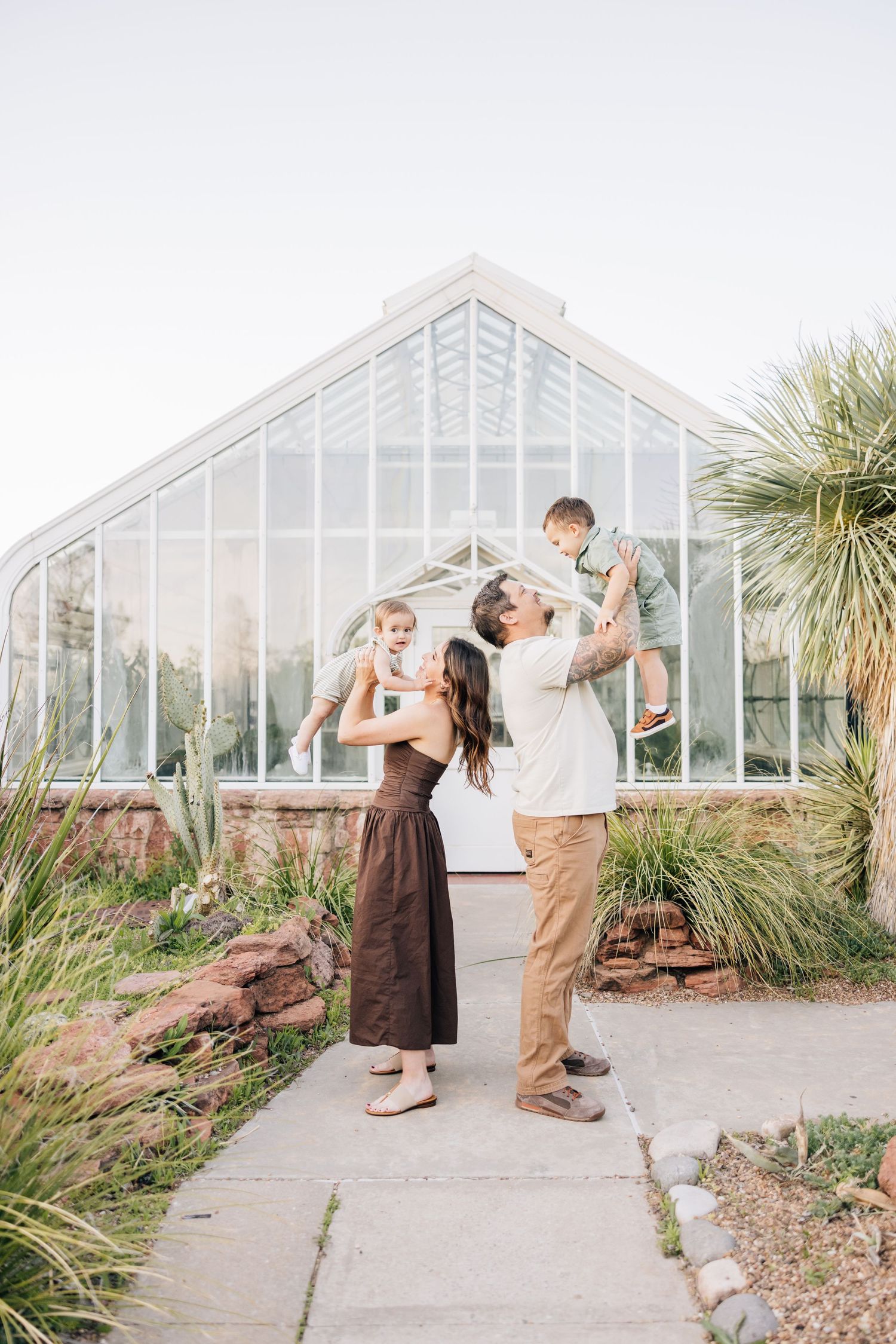 A couple stands with their children outside a modern greenhouse surrounded by tropical plants on a sunny day.
