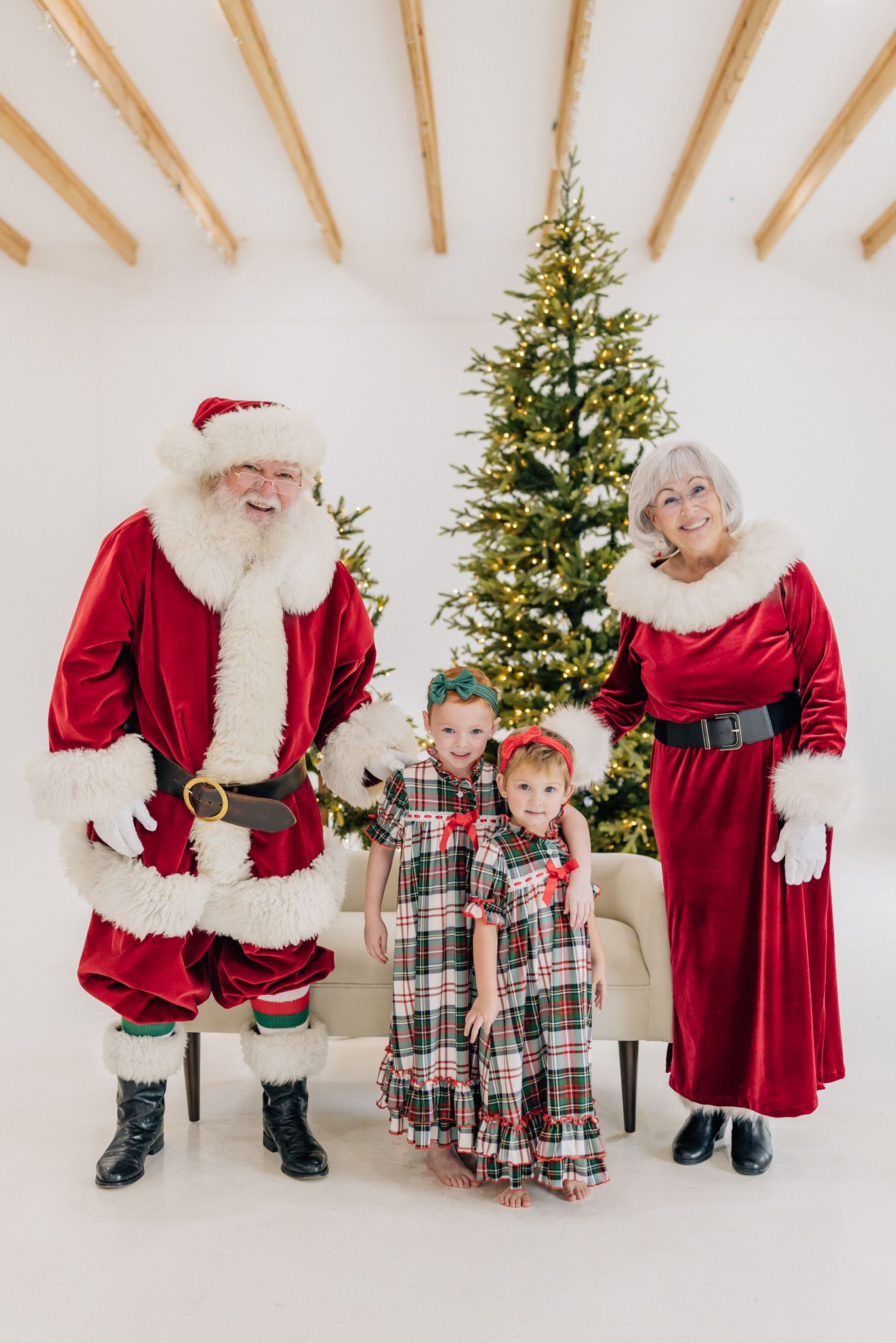 Two children in plaid pajamas pose with Santa and Mrs. Claus beside a decorated Christmas tree in a bright white studio.