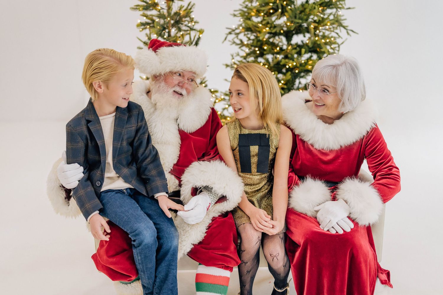 Two children sit with Santa Claus and Mrs. Claus in front of a decorated Christmas tree in a festive holiday photo.