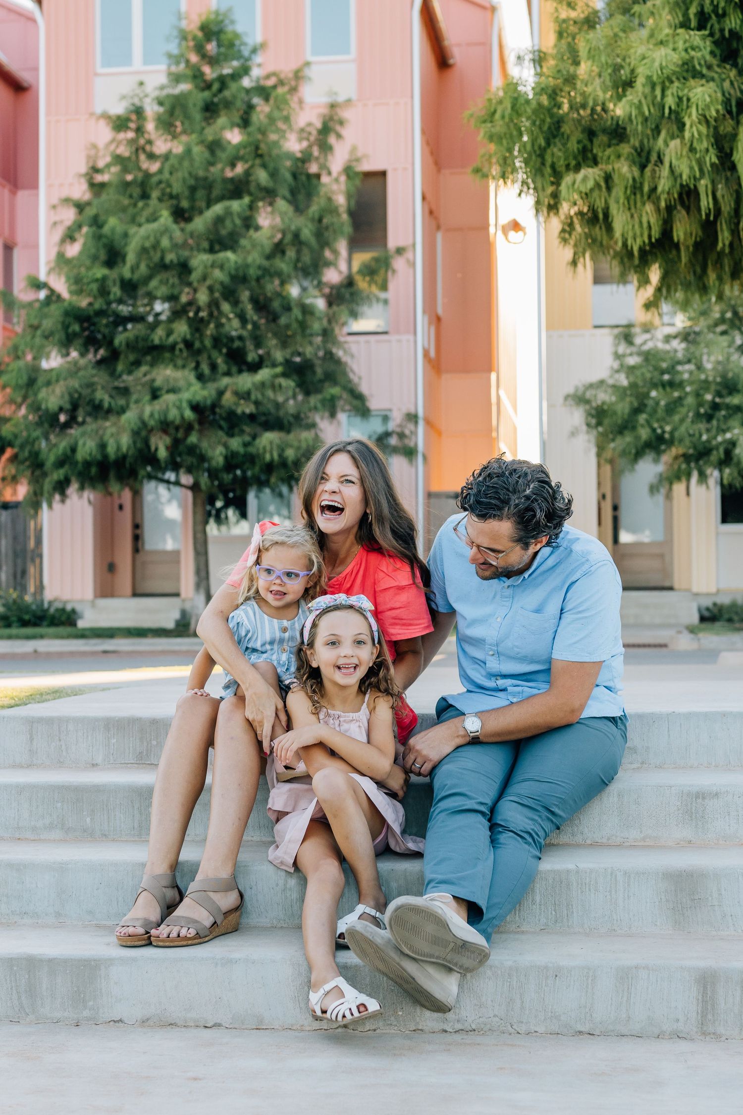 Family sharing joyful laughter while sitting on concrete steps near colorful buildings.