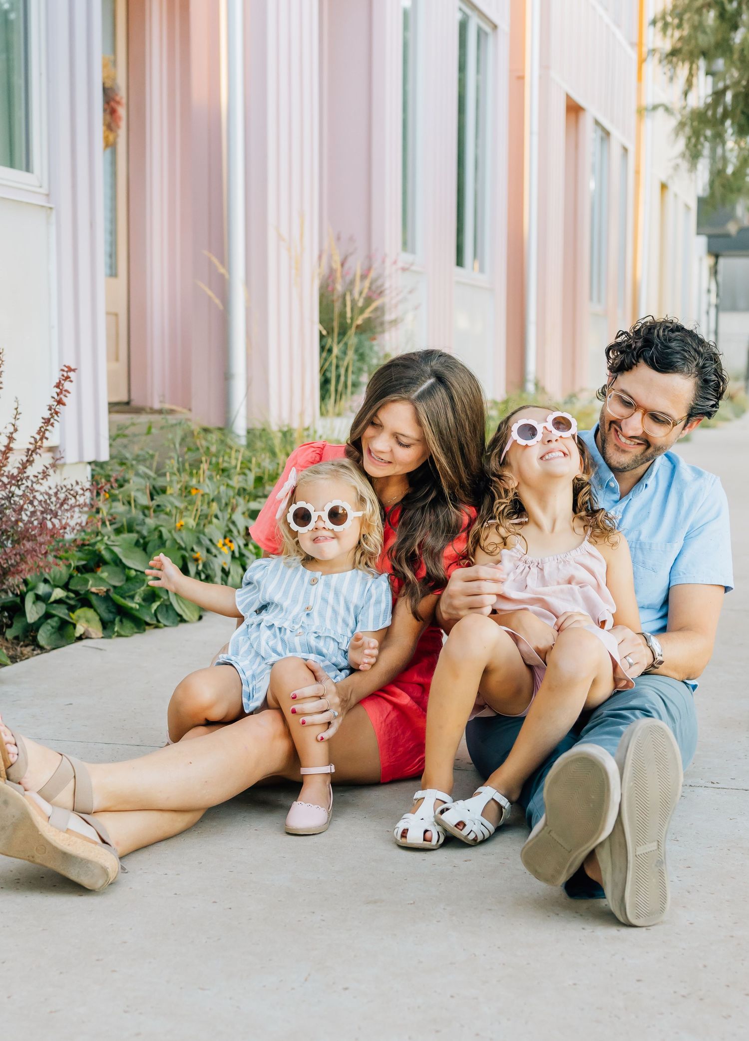 Family sitting together on sidewalk wearing sunglasses and casual outfits next to pink buildings and plants.