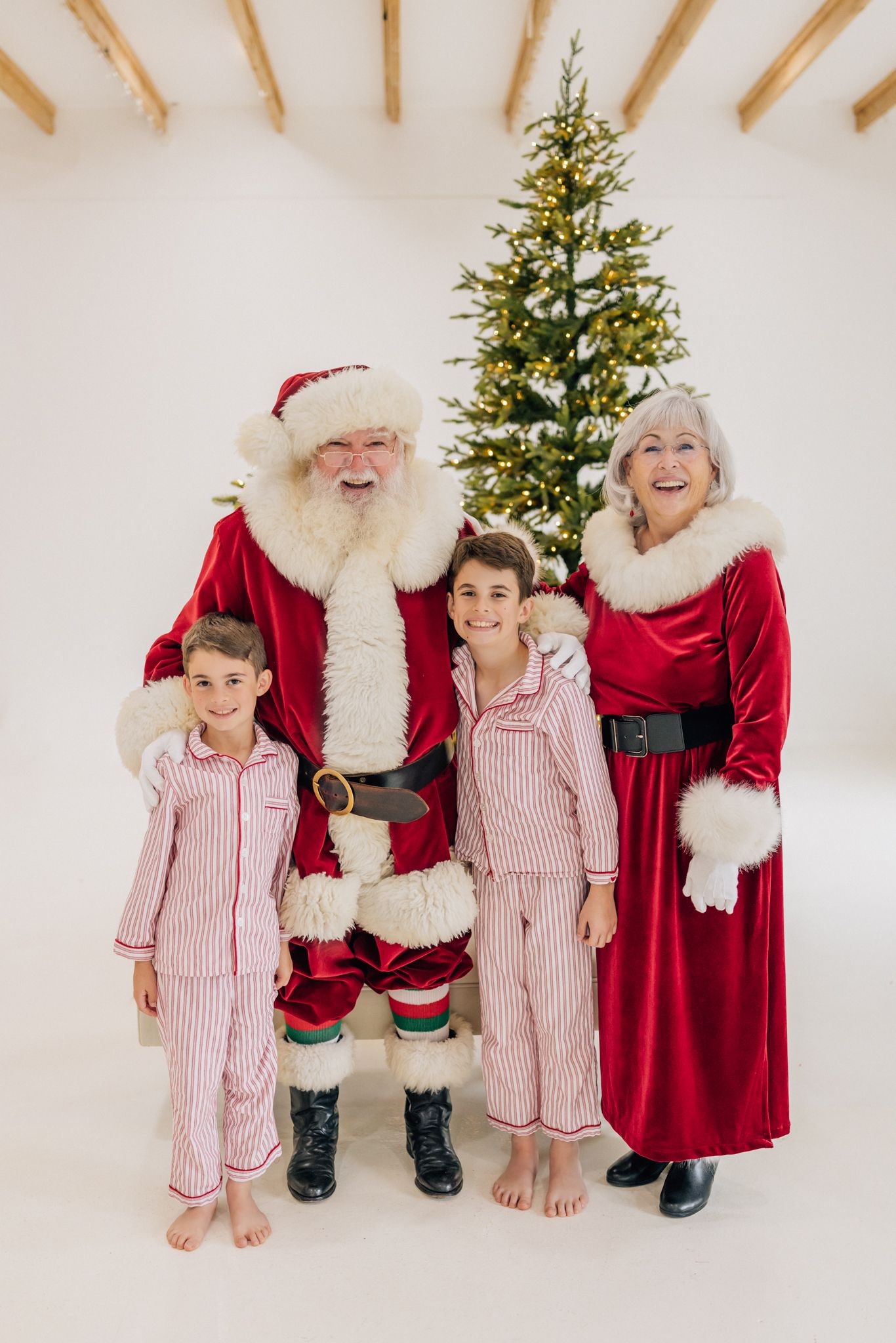 Santa and Mrs. Claus pose with two children in striped pajamas in front of a lit Christmas tree.