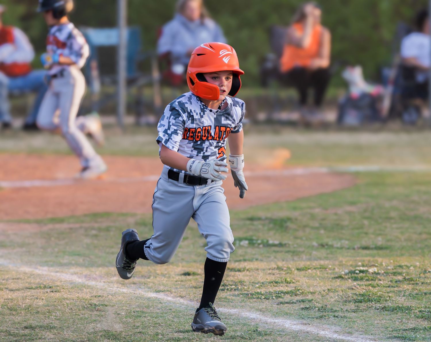 A youth baseball player in a gray uniform and red helmet runs down the first base line on a dirt baseball field.