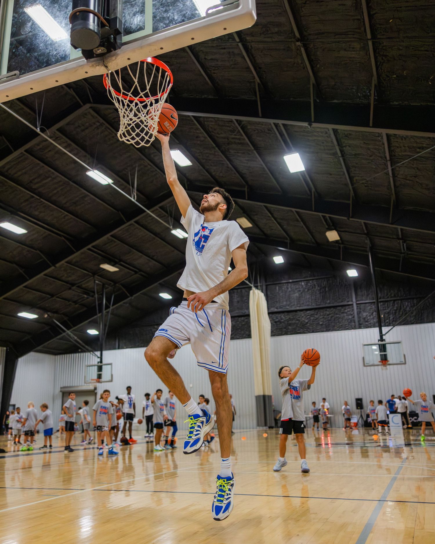 Basketball player in a white jersey leaps high for a layup during an indoor gym practice session.