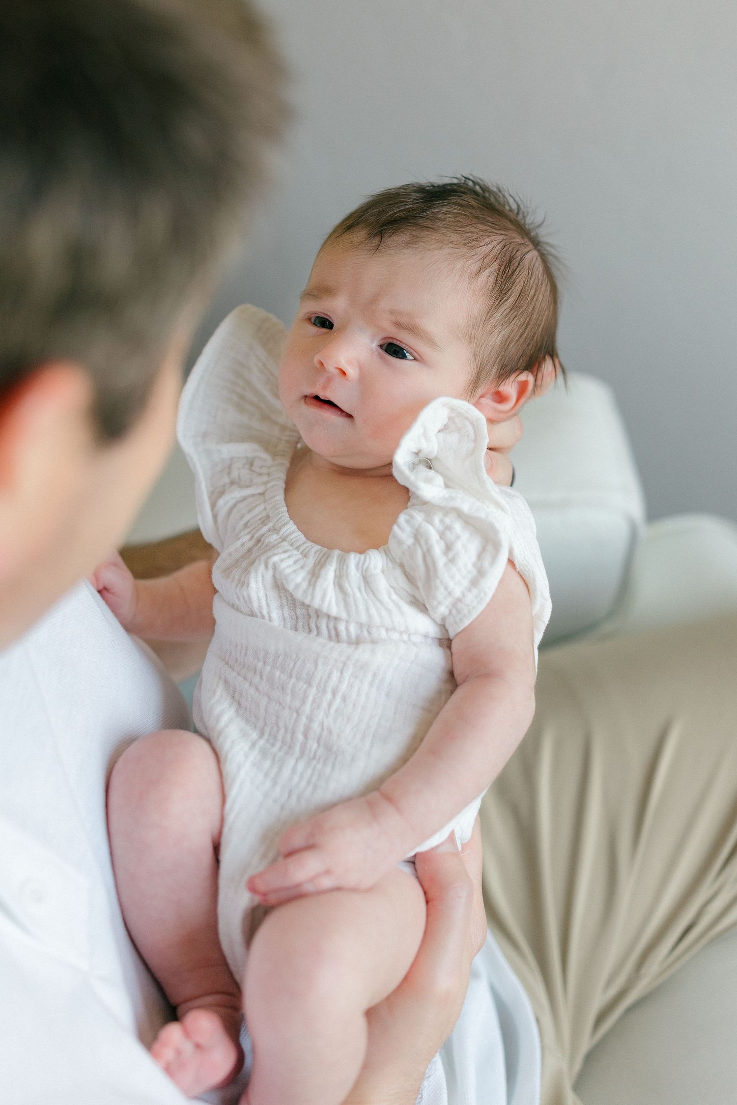 Father holding newborn baby girl in white ruffle onesie, infant looking up with curious expression.