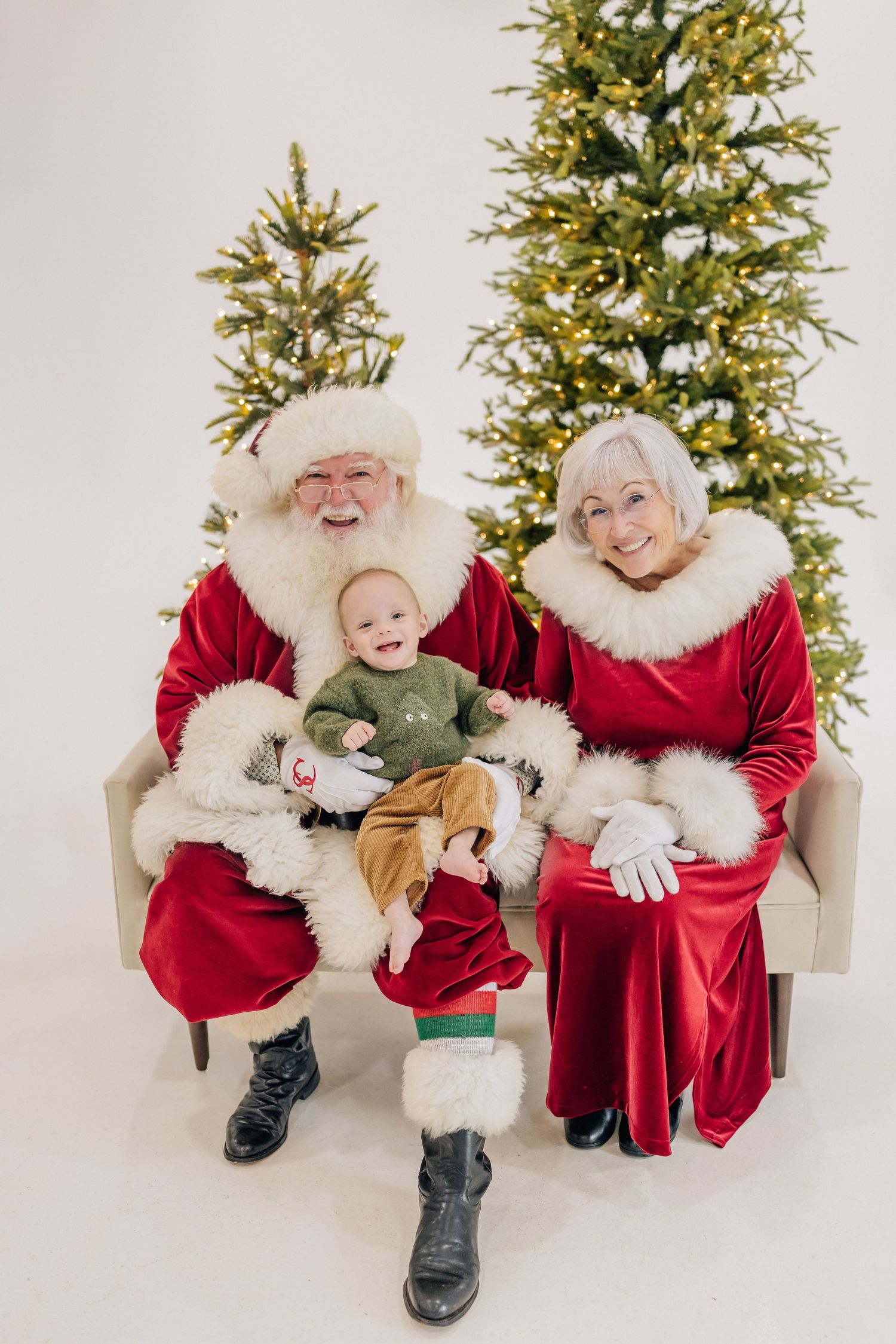 Santa and Mrs. Claus sit on a bench with a smiling baby, surrounded by lit Christmas trees in a white studio.