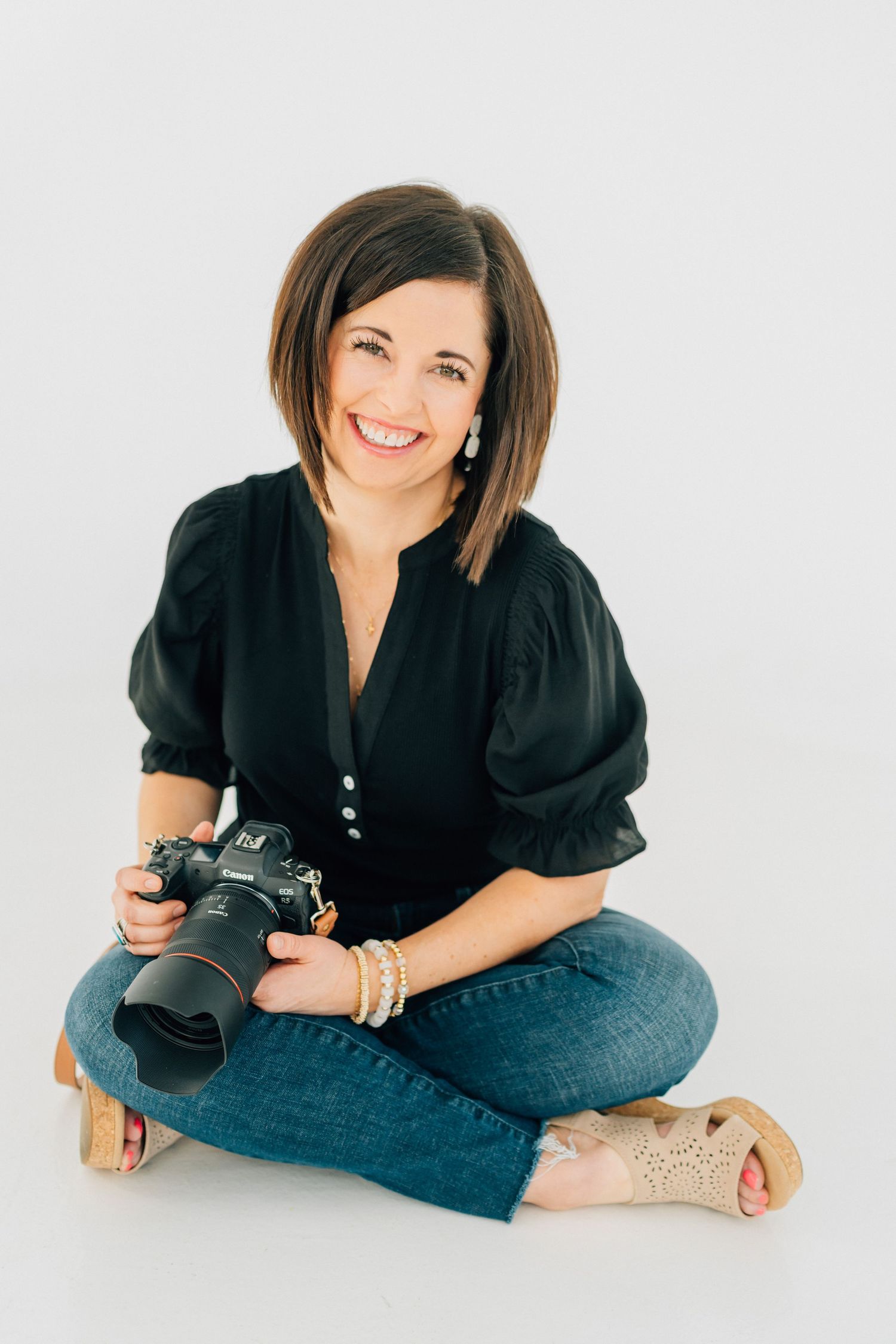 Smiling woman photographer sitting cross-legged on white background holding DSLR camera, wearing black blouse and jeans.