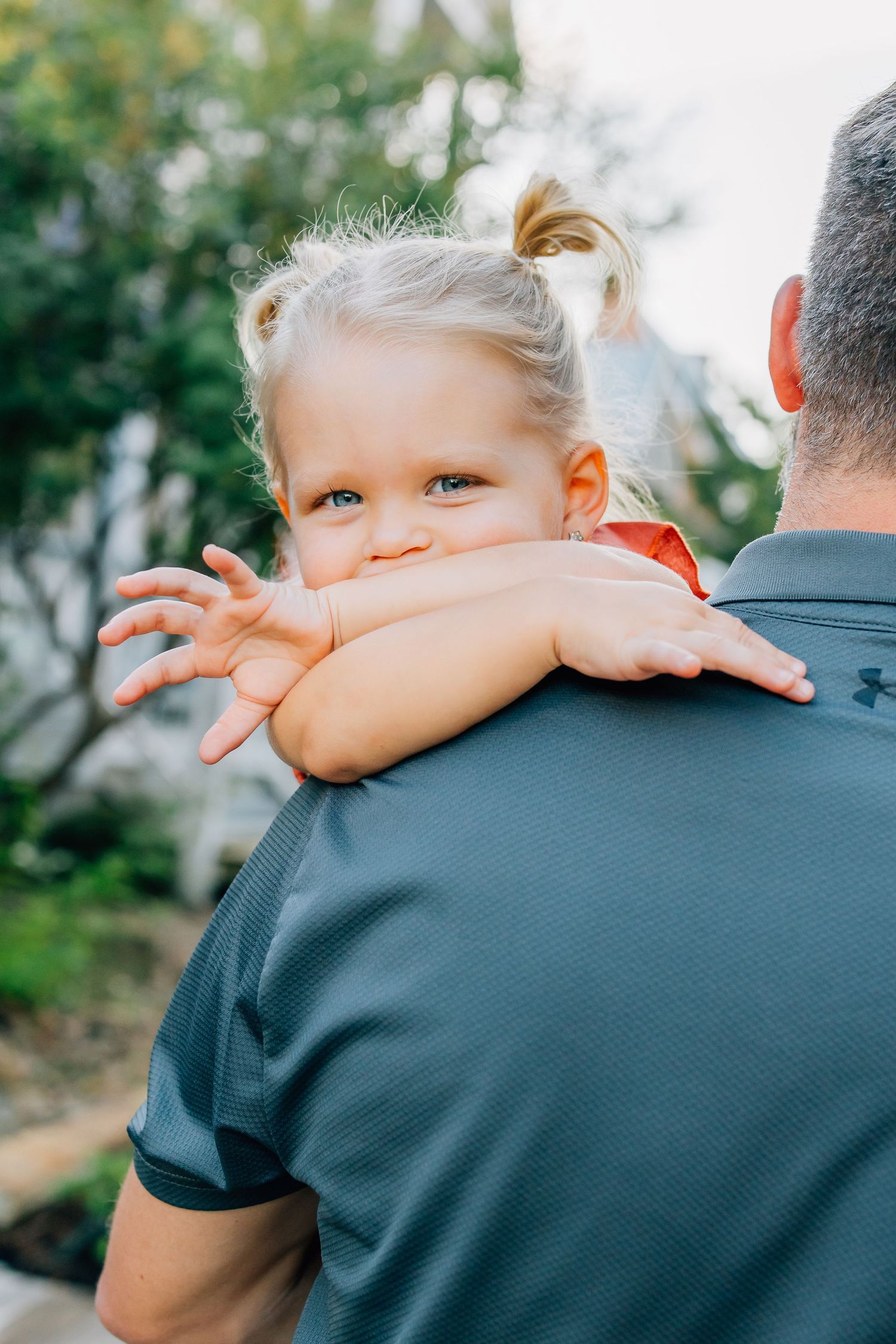 A small child with blonde hair peeks over an adult's shoulder while being carried outdoors on a sunny day.